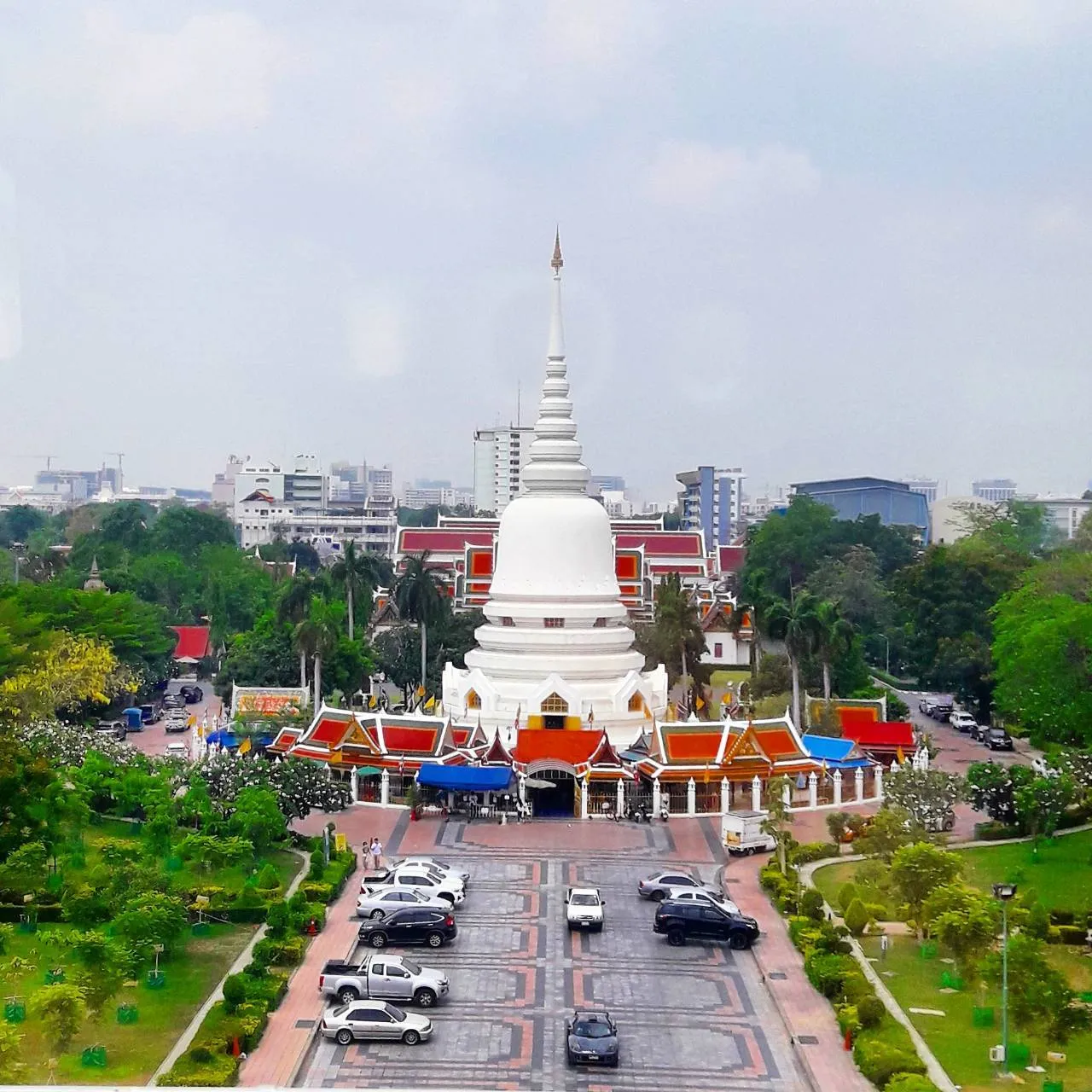 Wat Phra Si Mahathat in Bangkok seen from above, a tall white stupa with a slim spire standing behind red-roofed halls at the head of a broad paved approach lined with trees