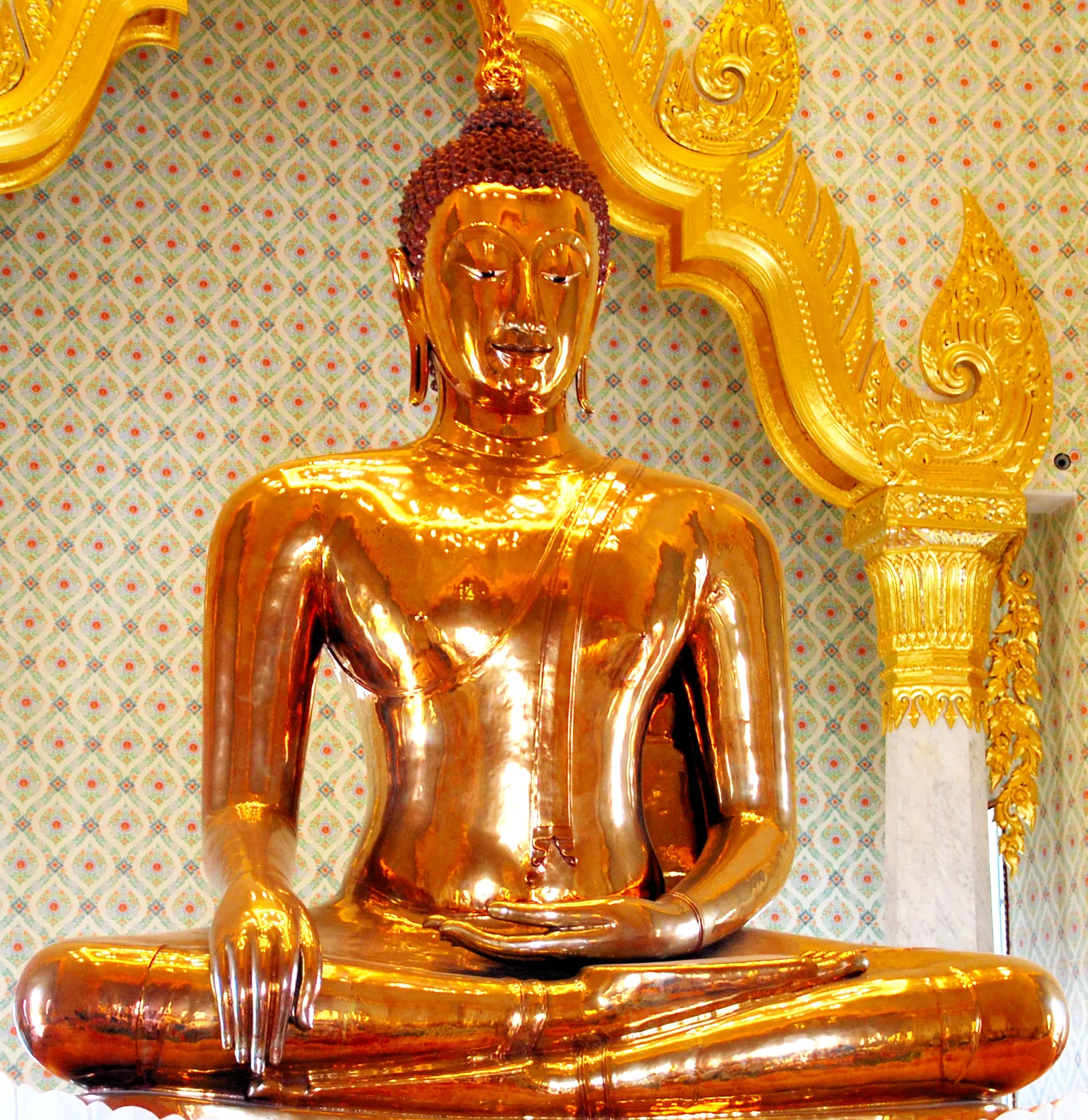 The Golden Buddha at Wat Traimit in Bangkok, a seated gold figure with one hand touching the ground, lit against a patterned wall beneath a gilded canopy