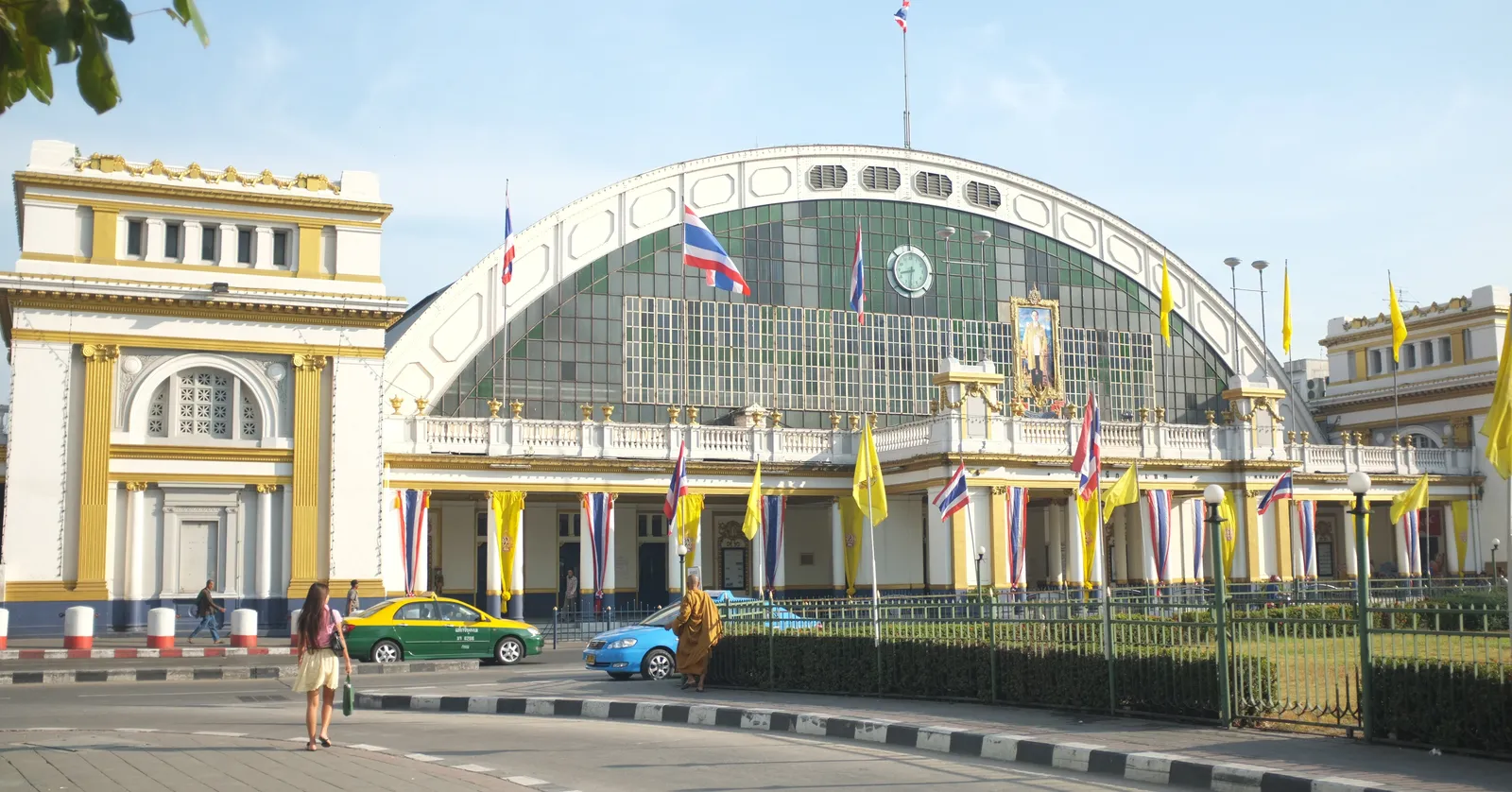 The front of Hua Lamphong station in Bangkok, a cream Italianate facade built around the semicircular end of an iron train shed, flags along the railings and taxis waiting at the kerb