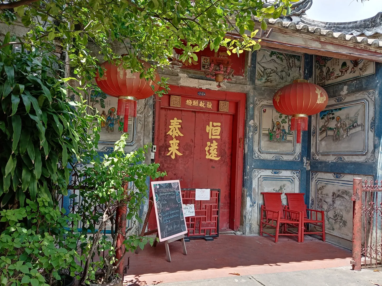 The entrance to So Heng Tai Mansion in Talat Noi, Bangkok, red-painted timber doors under gold Chinese lettering with paper lanterns hanging above and faded painted panels on the flanking walls
