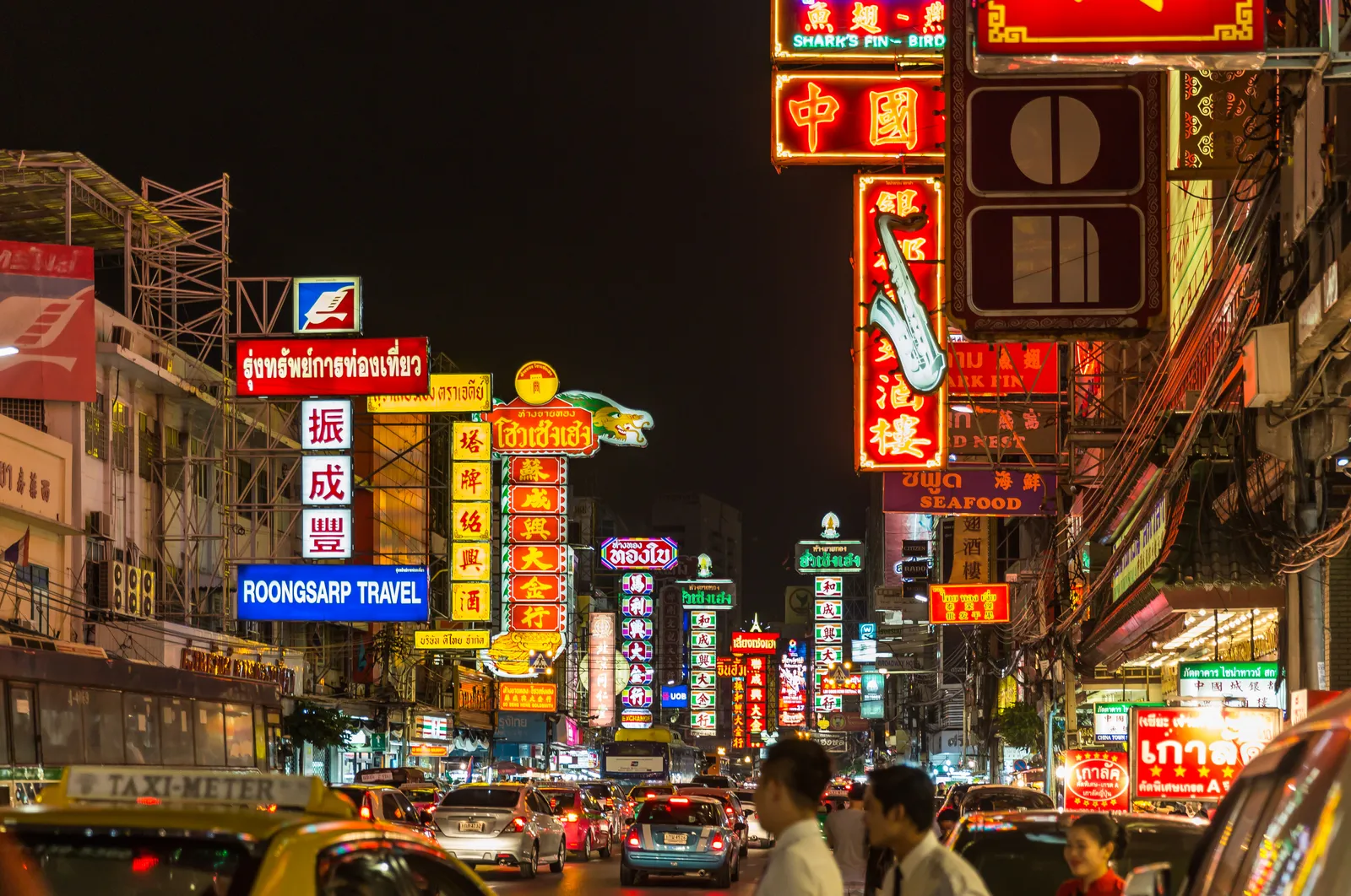 Yaowarat Road in Bangkok after dark, both sides of the street stacked with lit Chinese and Thai neon shop signs above slow traffic and people crossing between the stalls