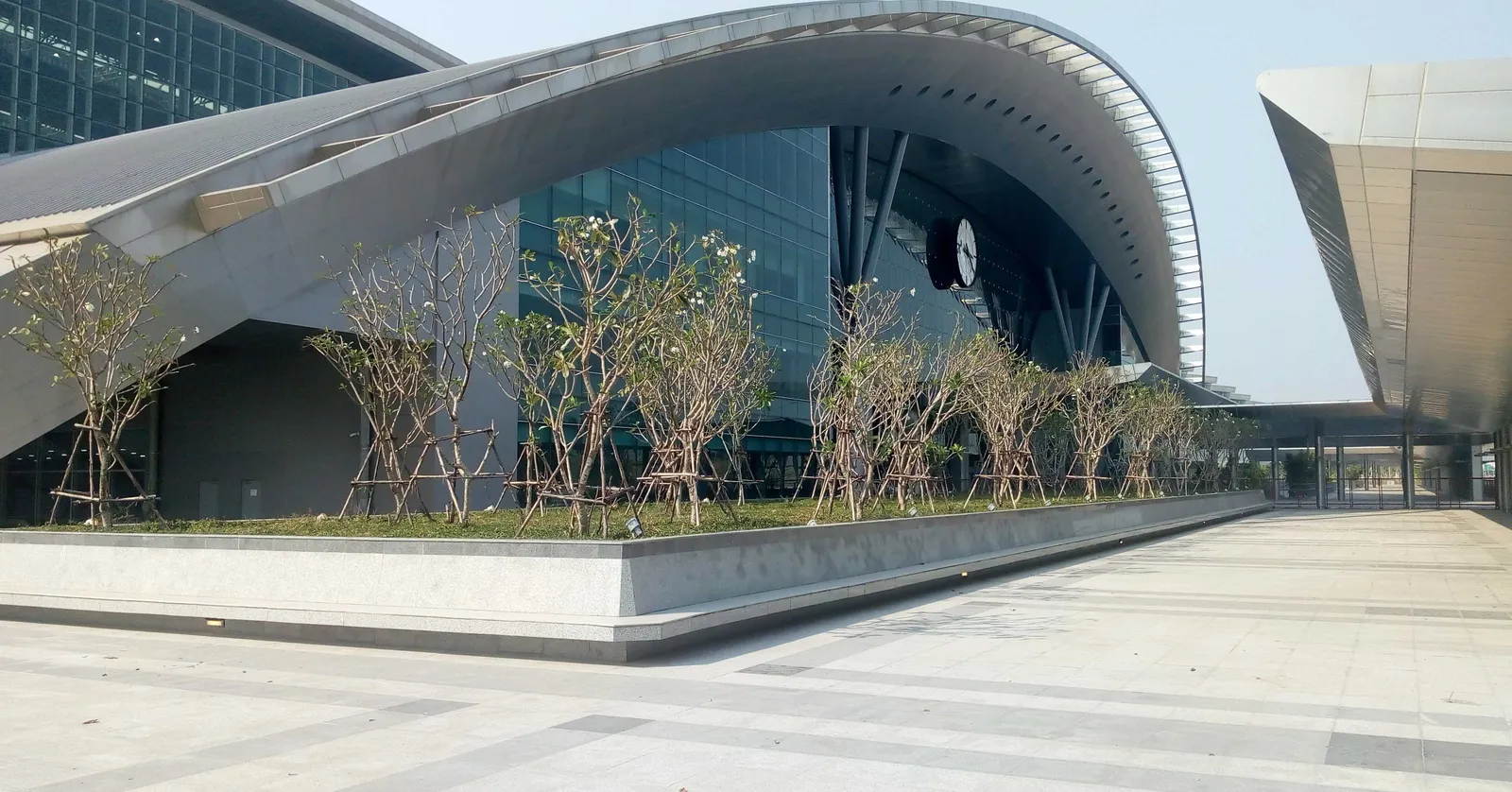 The exterior of Krung Thep Aphiwat Central Terminal in Bangkok, a long curved white roof sweeping over a glazed frontage above a wide empty plaza planted with young trees