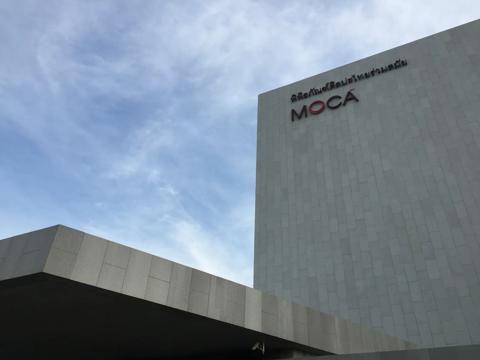 The pale stone exterior of the Museum of Contemporary Art in Bangkok, a tall windowless block carved with a repeating relief pattern, the museum's name mounted near the top corner against a clouded sky