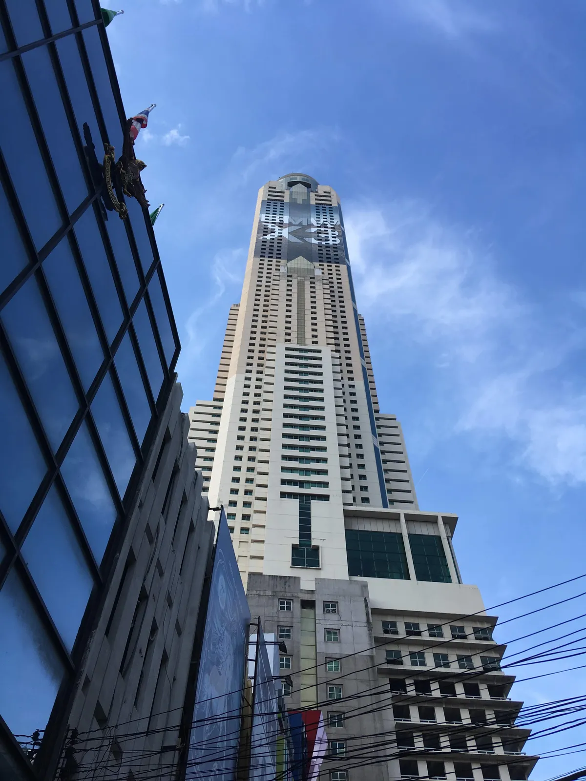 Baiyoke Tower II in Bangkok seen from street level, a pale tapering high-rise rising far above its neighbours with a dark glass band and a mast at the very top