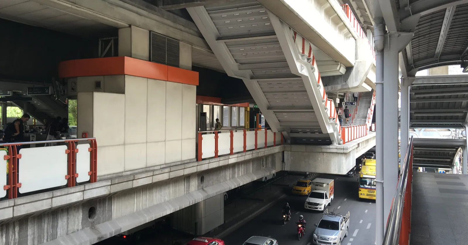 Phaya Thai station in Bangkok seen from the side, a concrete elevated platform box and its linking walkway carried over a street of cars and trucks on heavy piers