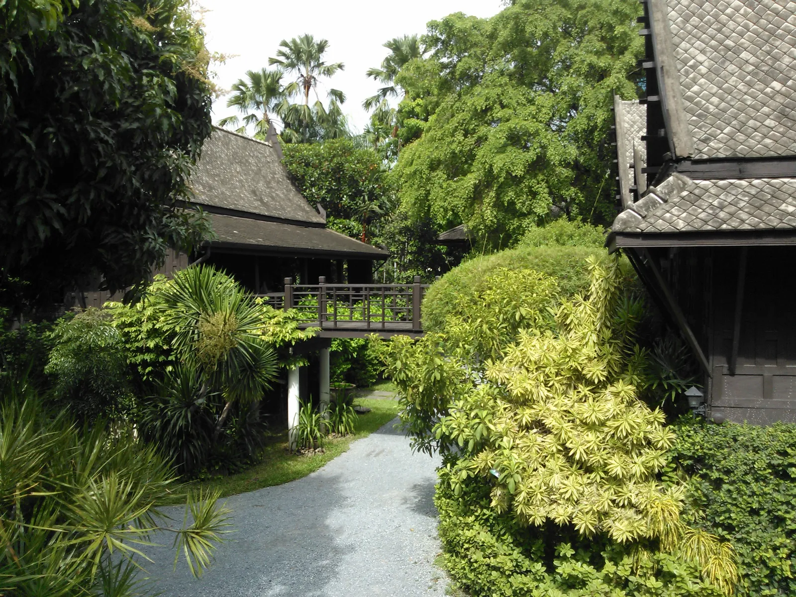 Traditional Thai teak houses with steep dark tiled roofs and a raised connecting walkway at Suan Pakkad Palace in Bangkok, set among palms and clipped shrubs along a gravel path