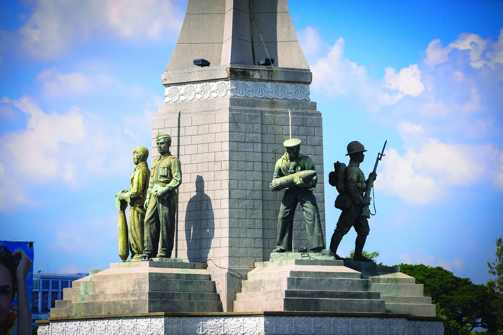 The Victory Monument in Bangkok, a tall grey obelisk of clasped bayonet shapes on a circular plinth in the middle of a traffic roundabout, with buses and taxis circling it under an elevated railway