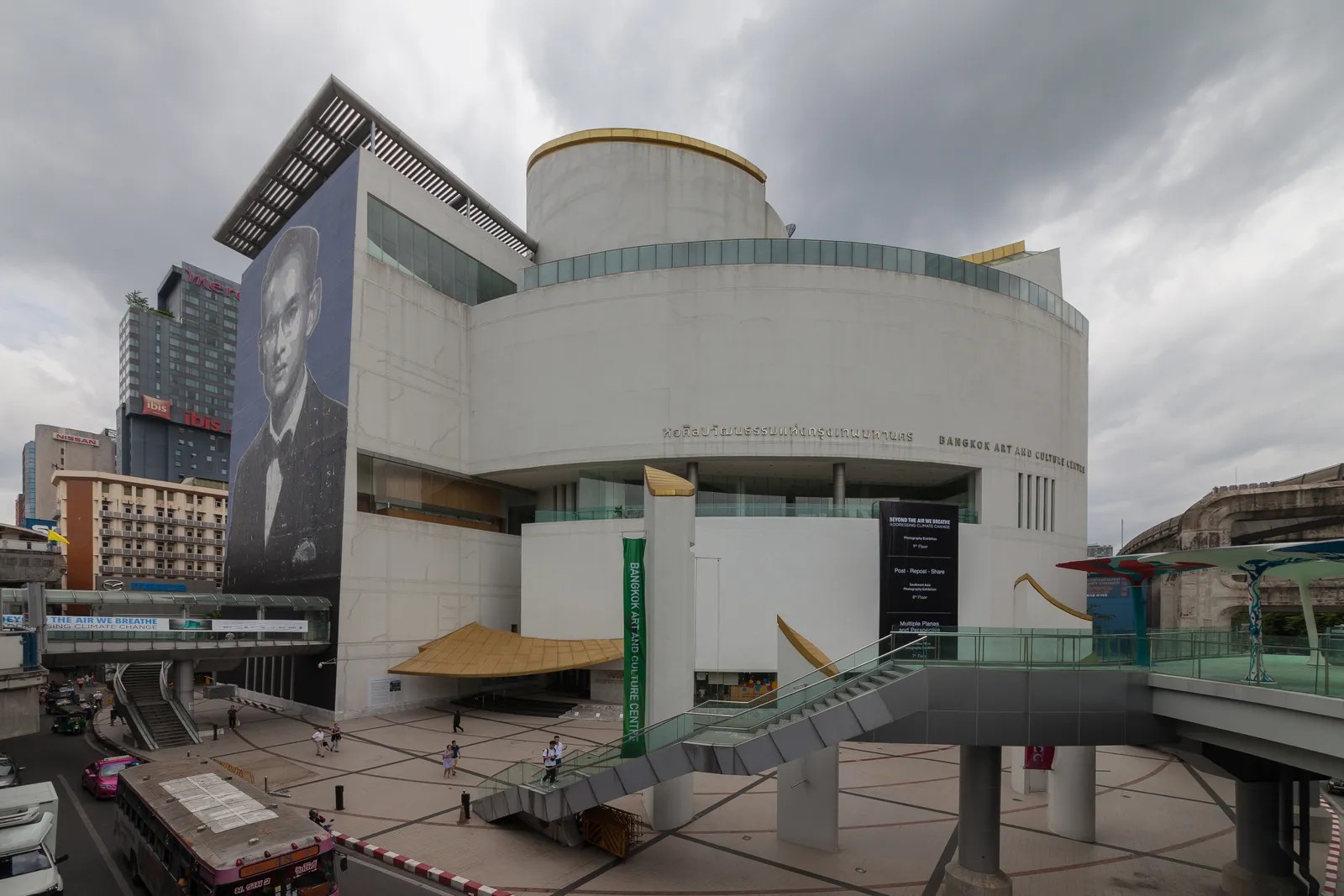 The Bangkok Art and Culture Centre, a white curved building with a drum-shaped upper storey standing over a plaza, with an elevated walkway running to its entrance from the right
