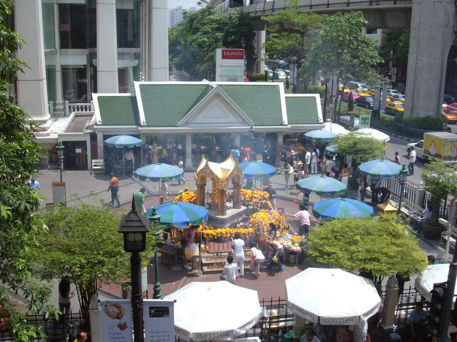 The Erawan Shrine in Bangkok seen from the walkway above it, a small gilded pavilion heaped with marigold garlands on a street corner, surrounded by blue and white umbrellas and people making offerings