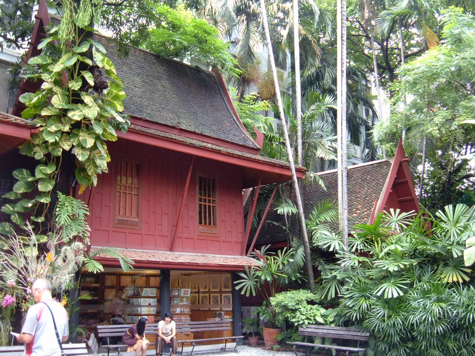 A dark red teak house at the Jim Thompson House in Bangkok, raised on posts under a steep tiled roof with carved gable finials, hemmed in by palms and hanging ferns