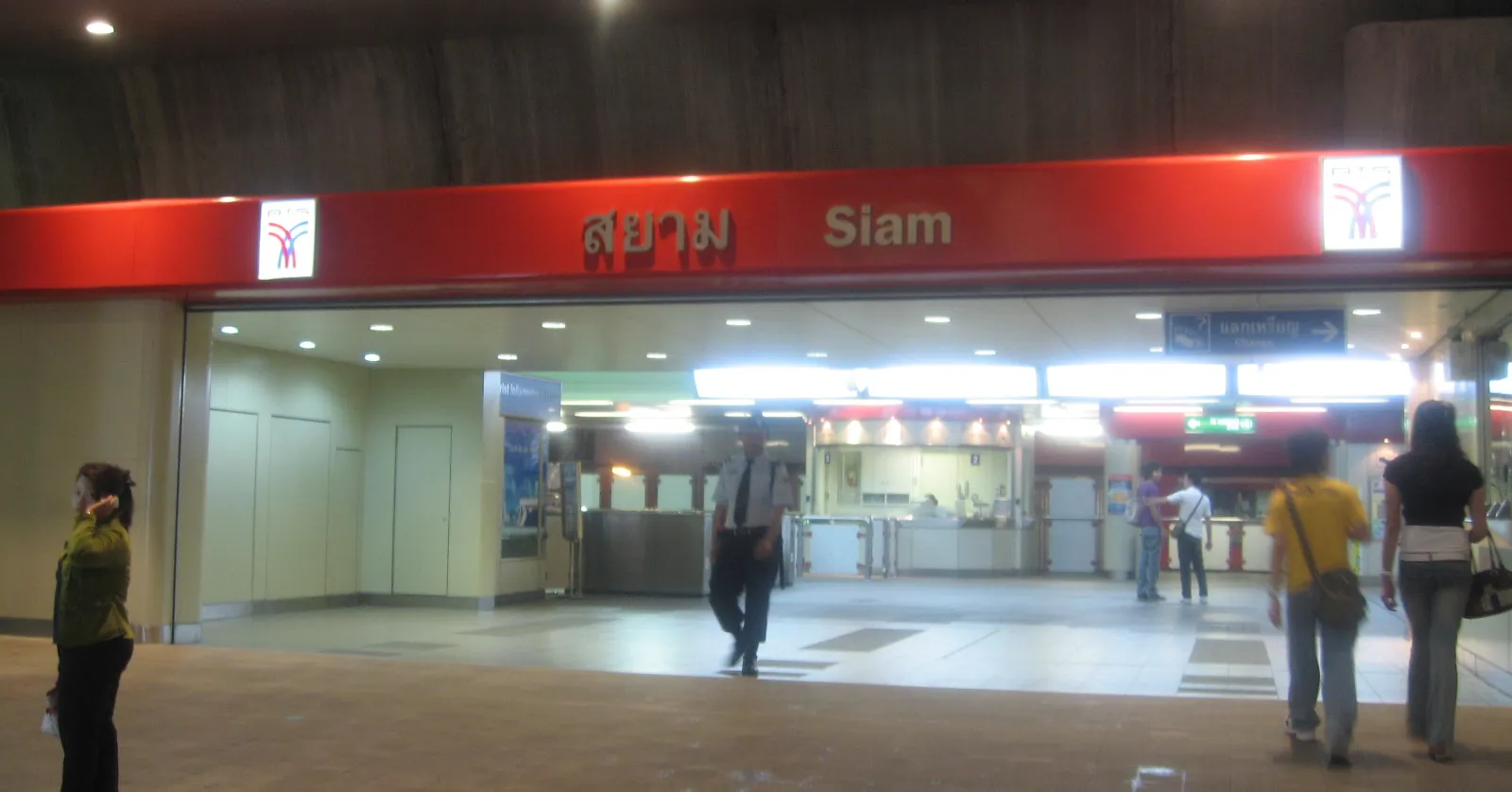 The concourse level of Siam station in Bangkok, a red-panelled ticket hall under the elevated tracks with the station name in Thai and English above the gateline and passengers crossing in front