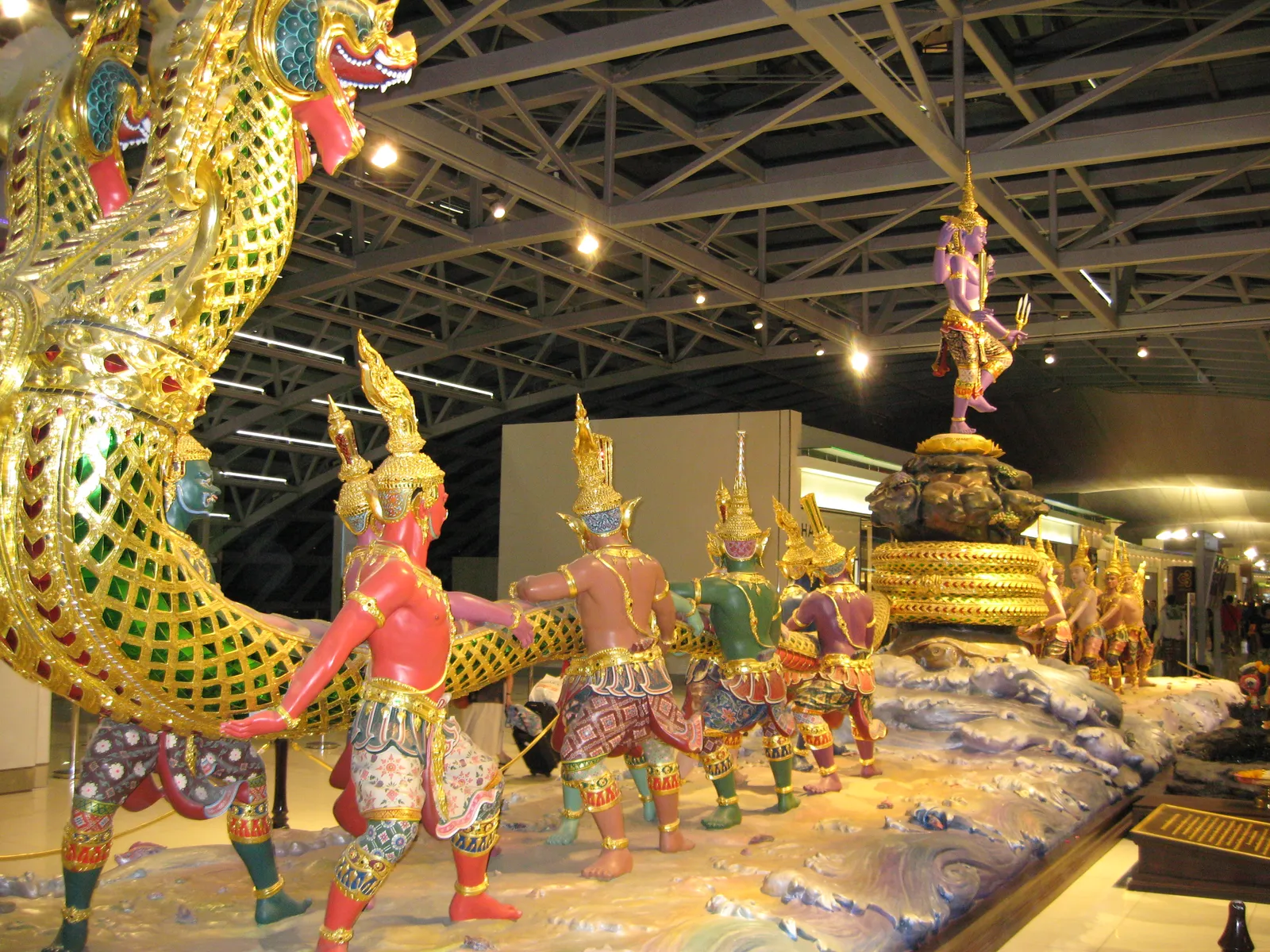 The Churning of the Ocean of Milk sculpture at Suvarnabhumi Airport, a long green and gold mirror-inlaid serpent hauled by ranks of painted gods and demons over modelled waves, with a gilded figure standing on a mountain at the far end