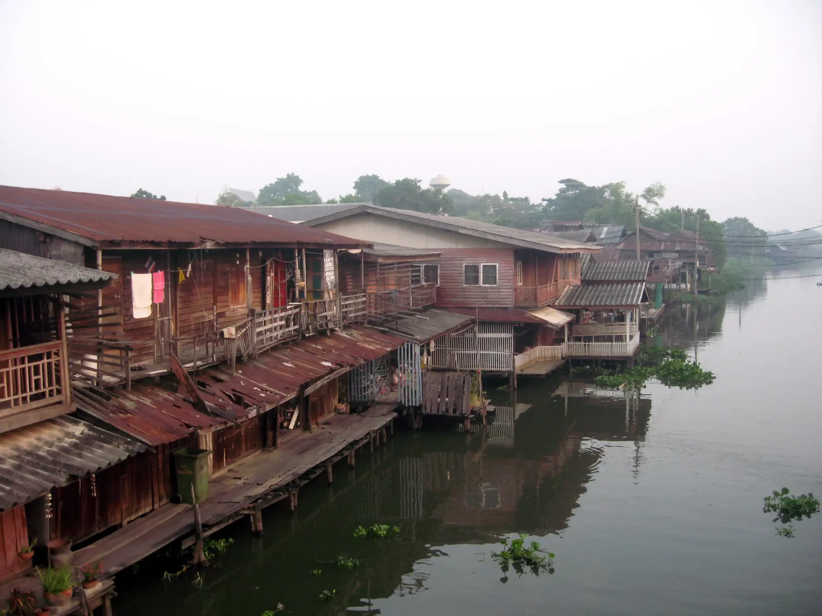 A row of old timber houses on stilts along the canal at Hua Takhe in Bangkok, rusted corrugated roofs and weathered plank walls stepping down to the brown water with washing hung on the balconies
