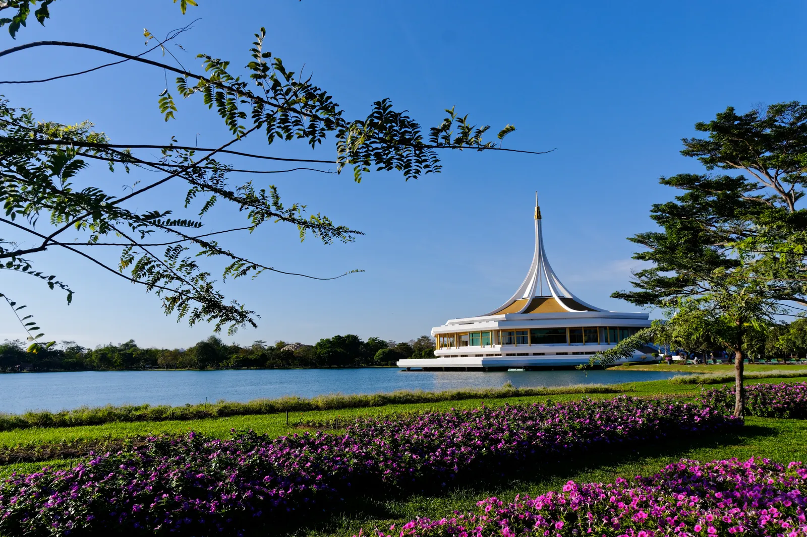 The Chai Chon pavilion at Suan Luang Rama IX park in Bangkok, a white building under a tall pointed golden-edged spire, seen across the lake from a bank planted with purple and pink flower beds