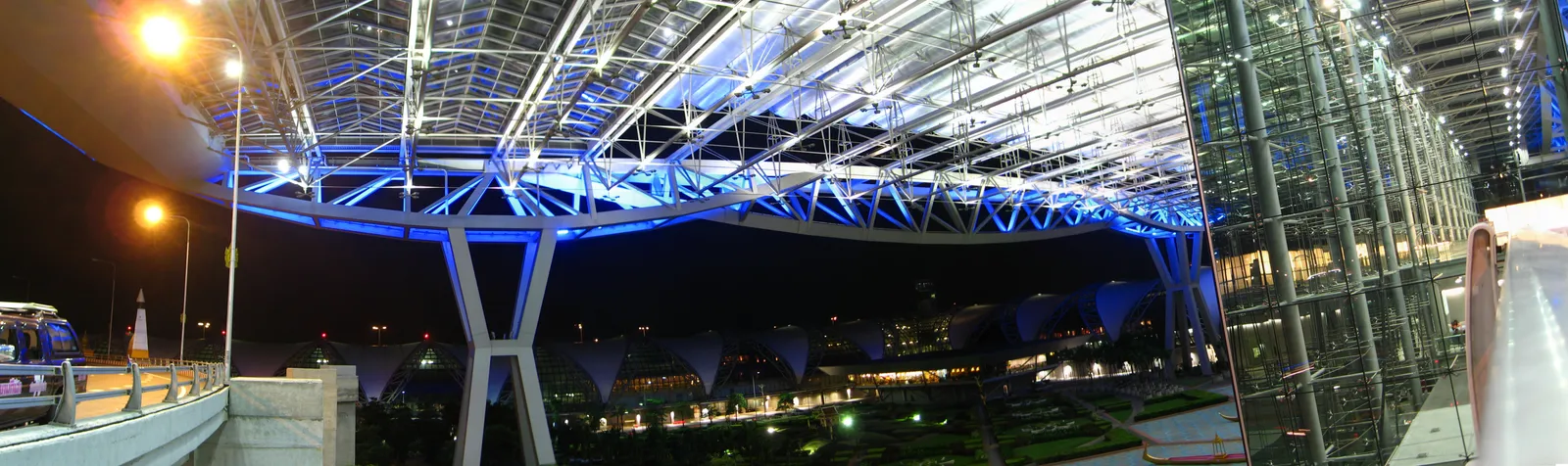 The cantilevered roof of Suvarnabhumi Airport's terminal lit blue and white at night, its steel trusses reaching out over the landscaped forecourt below