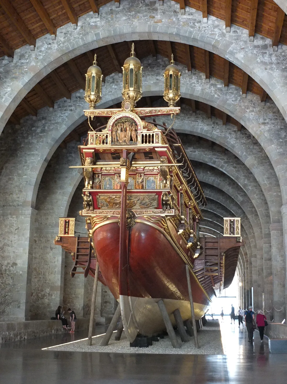 The full-size replica of the Galera Real, a red and gilded sixteenth-century royal galley with three lanterns on its stern, under the stone arches of the medieval shipyards at the Maritime Museum of Barcelona