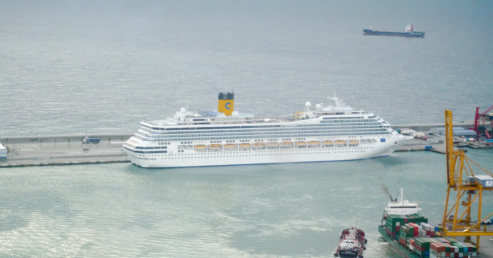 A white cruise ship with a yellow funnel tied up at the Adossat wharf in the Port of Barcelona, seen from Montjuïc, with a container ship and cranes in the foreground