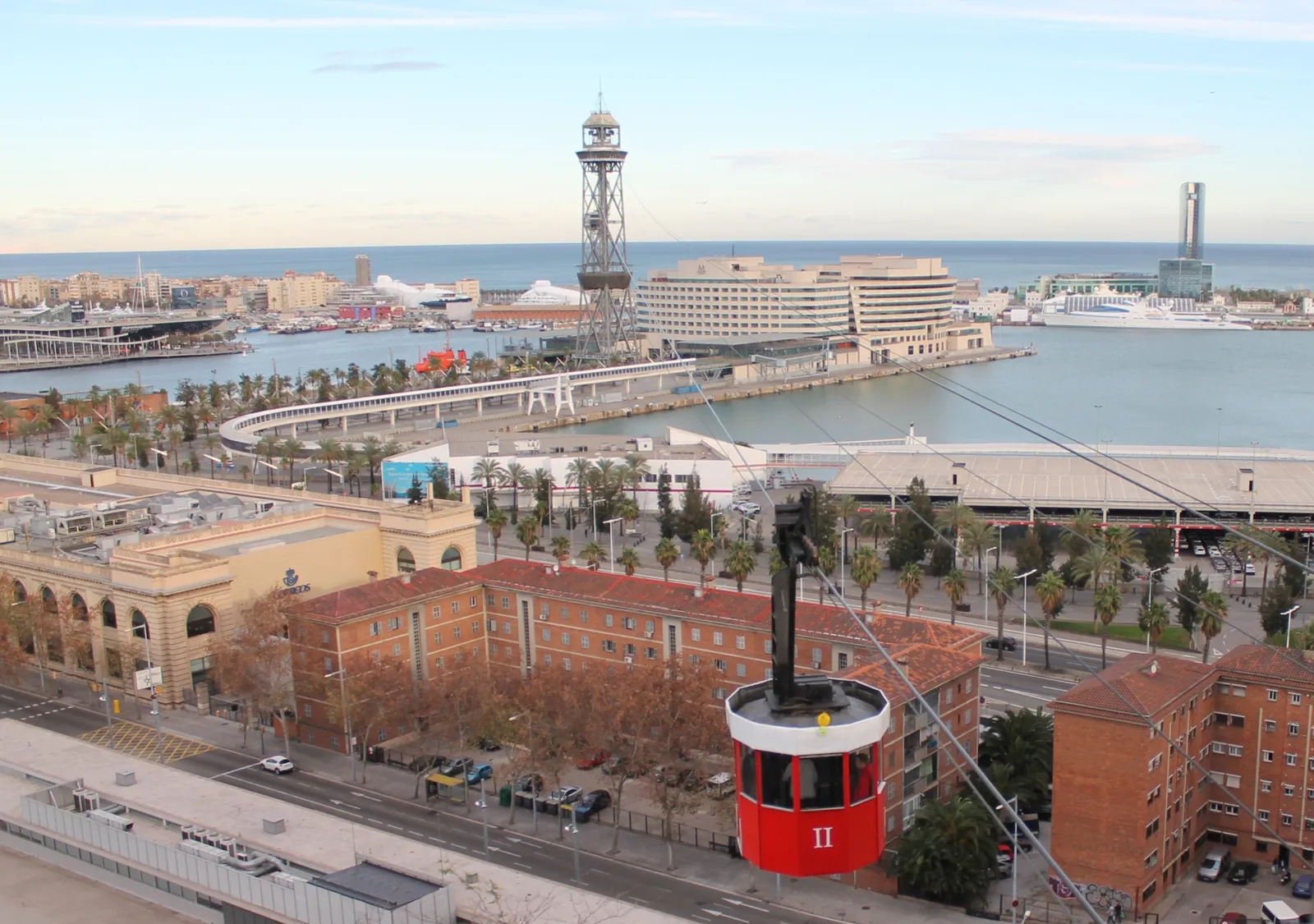 A red cabin of Barcelona's port cable car hanging over the old harbor, with the steel Torre de Jaume I, the World Trade Center buildings and cruise ships behind it