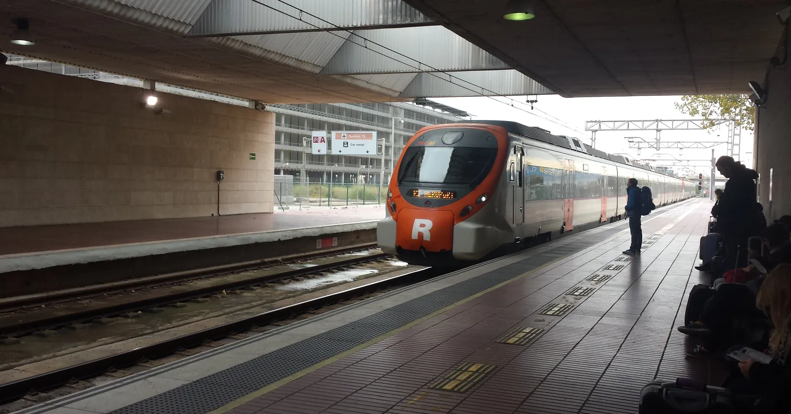 A red and white R2 Nord commuter train at the platform of the Aeroport railway station at Barcelona-El Prat airport, with passengers and luggage waiting