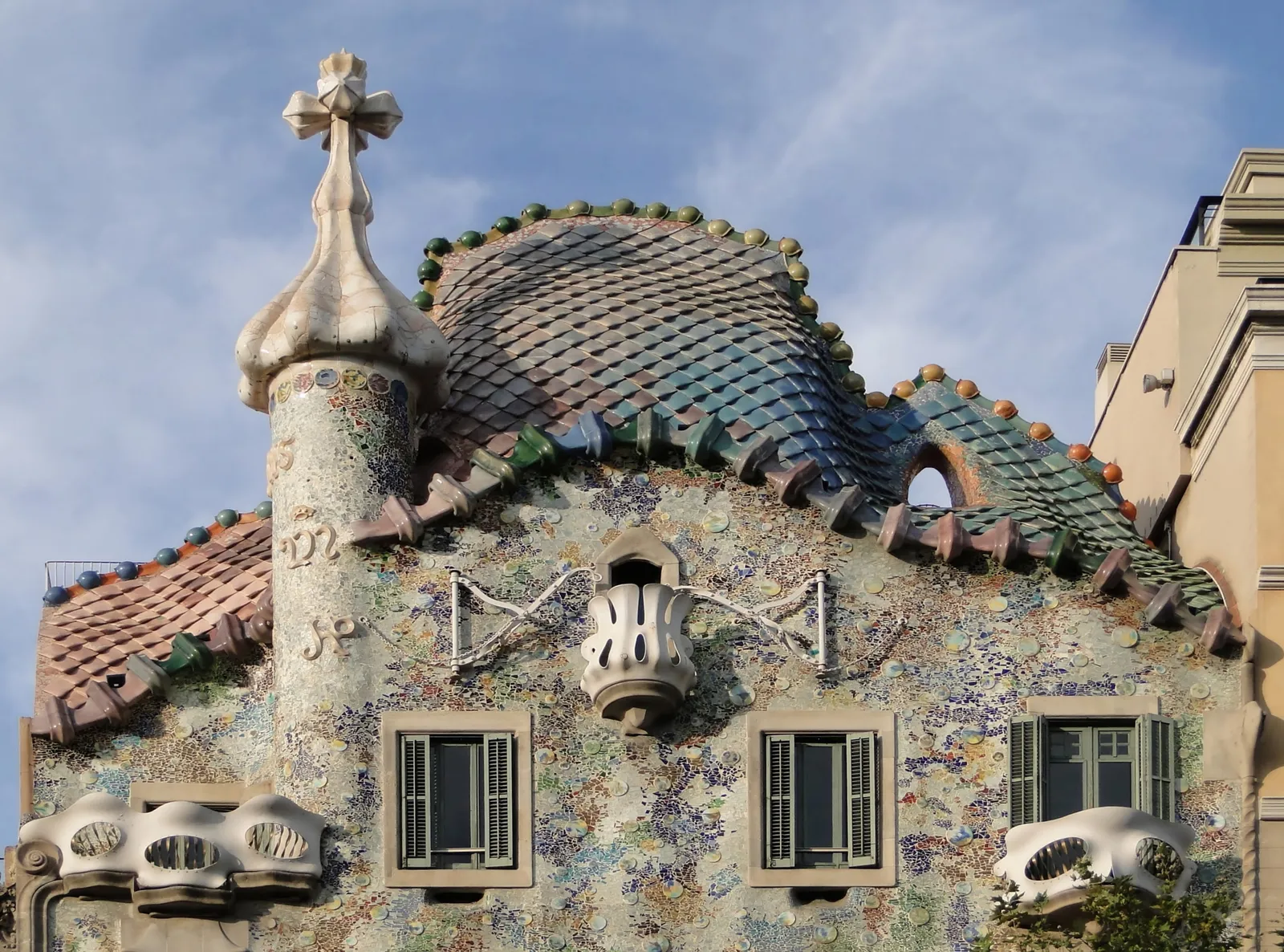 The top of Casa Batlló on Passeig de Gràcia in Barcelona, with a roof of scale-like tiles shaped like a dragon's back, a cross-topped turret and a mosaic front