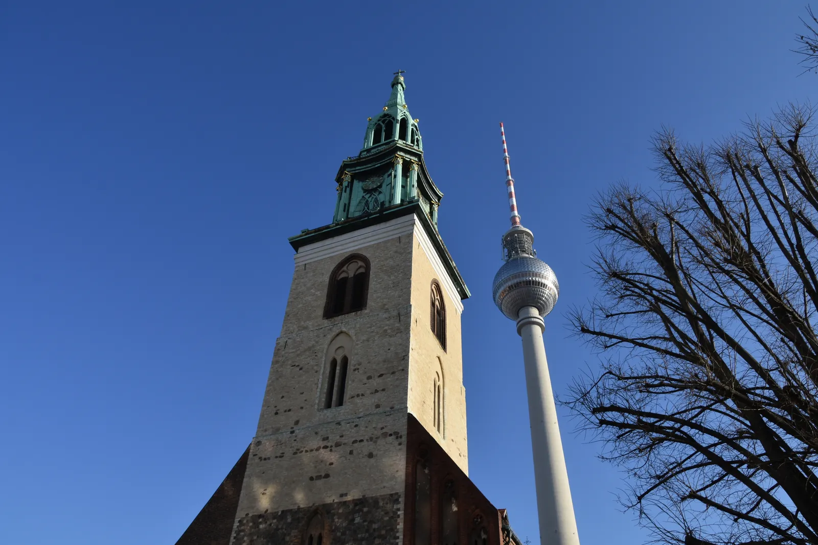 The stone tower and green copper spire of the Marienkirche next to the striped shaft and mirrored sphere of the Berlin TV Tower, both against a deep blue winter sky