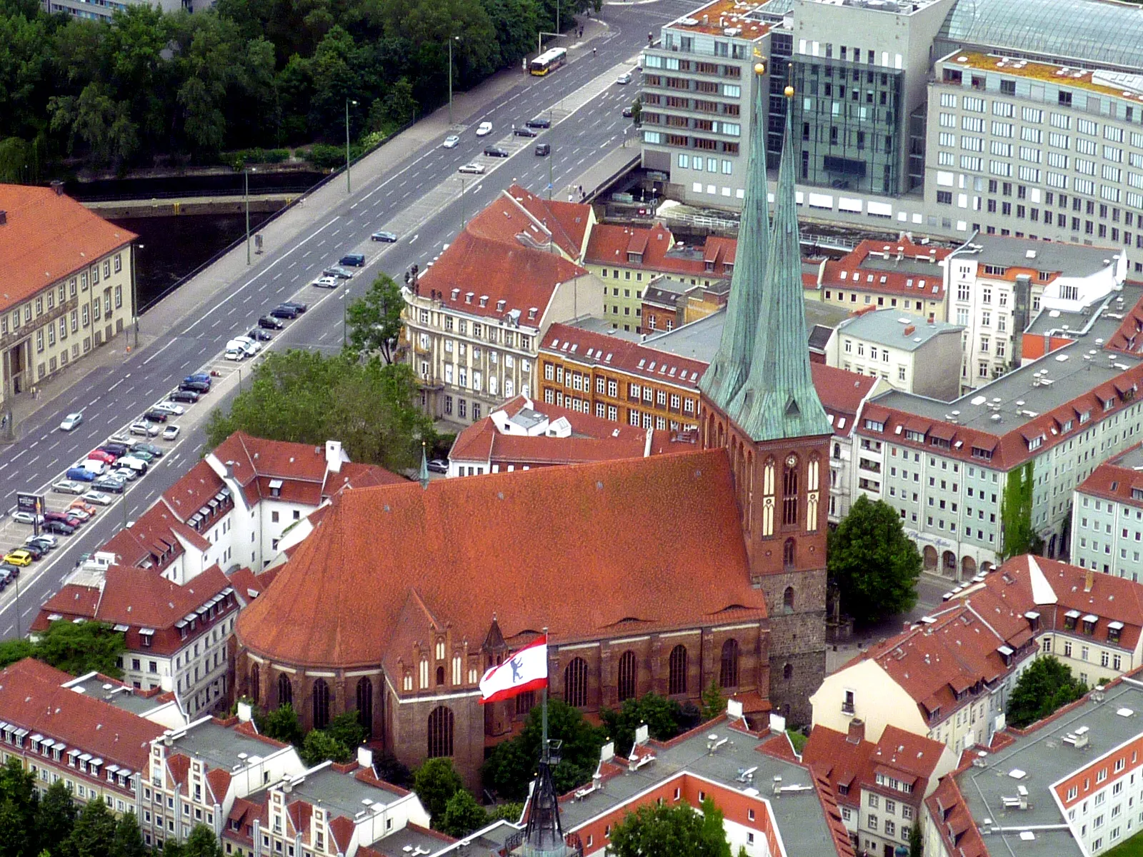 The Nikolaiviertel seen from above, the twin green spires of the Nikolaikirche rising from a dense block of red-tiled roofs beside the Spree, with modern Berlin streets behind