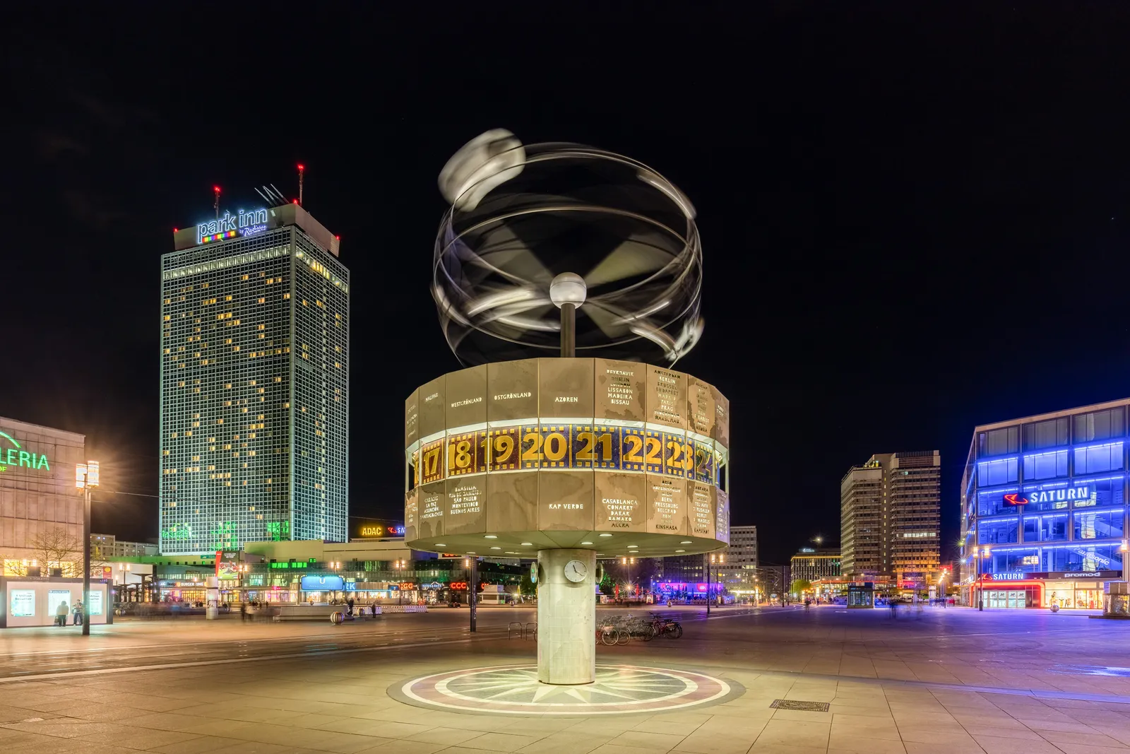 The Urania World Clock on Alexanderplatz at night, a lit rotating drum of city names and hour numbers under a metal model of the solar system, with the tower of the Park Inn hotel behind it