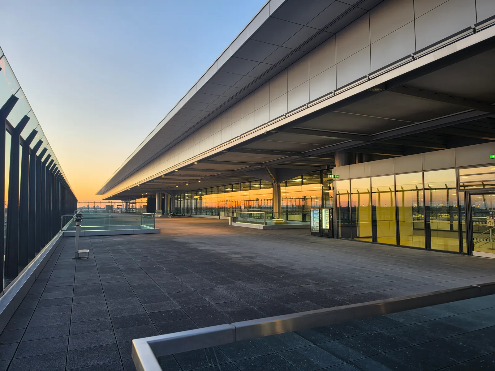 The open rooftop observation deck on top of Terminal 1 at Berlin Brandenburg Airport at sunset, with the terminal's long cantilevered roof running away to the left and the apron visible beyond the railing