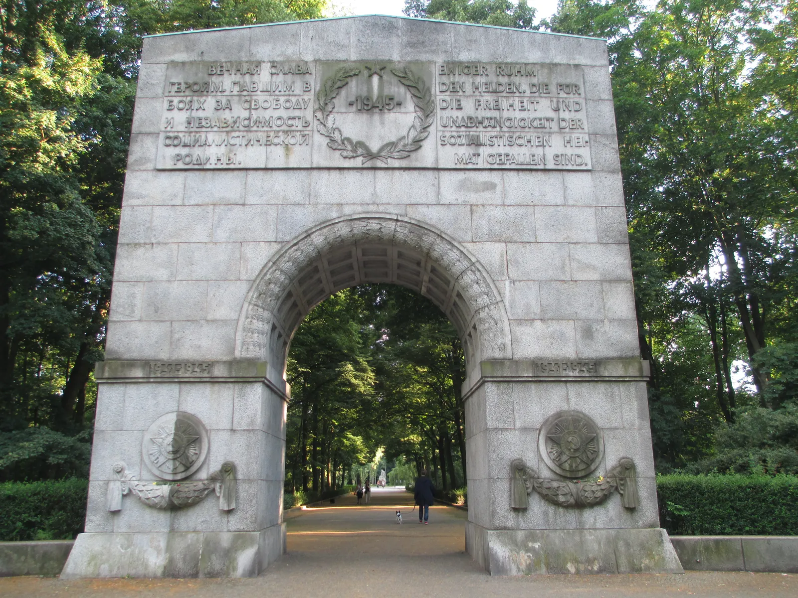 The grey granite entrance arch to the Soviet War Memorial in Treptower Park, Berlin, carved with Russian and German inscriptions, opening onto a tree-lined avenue with a few people walking through