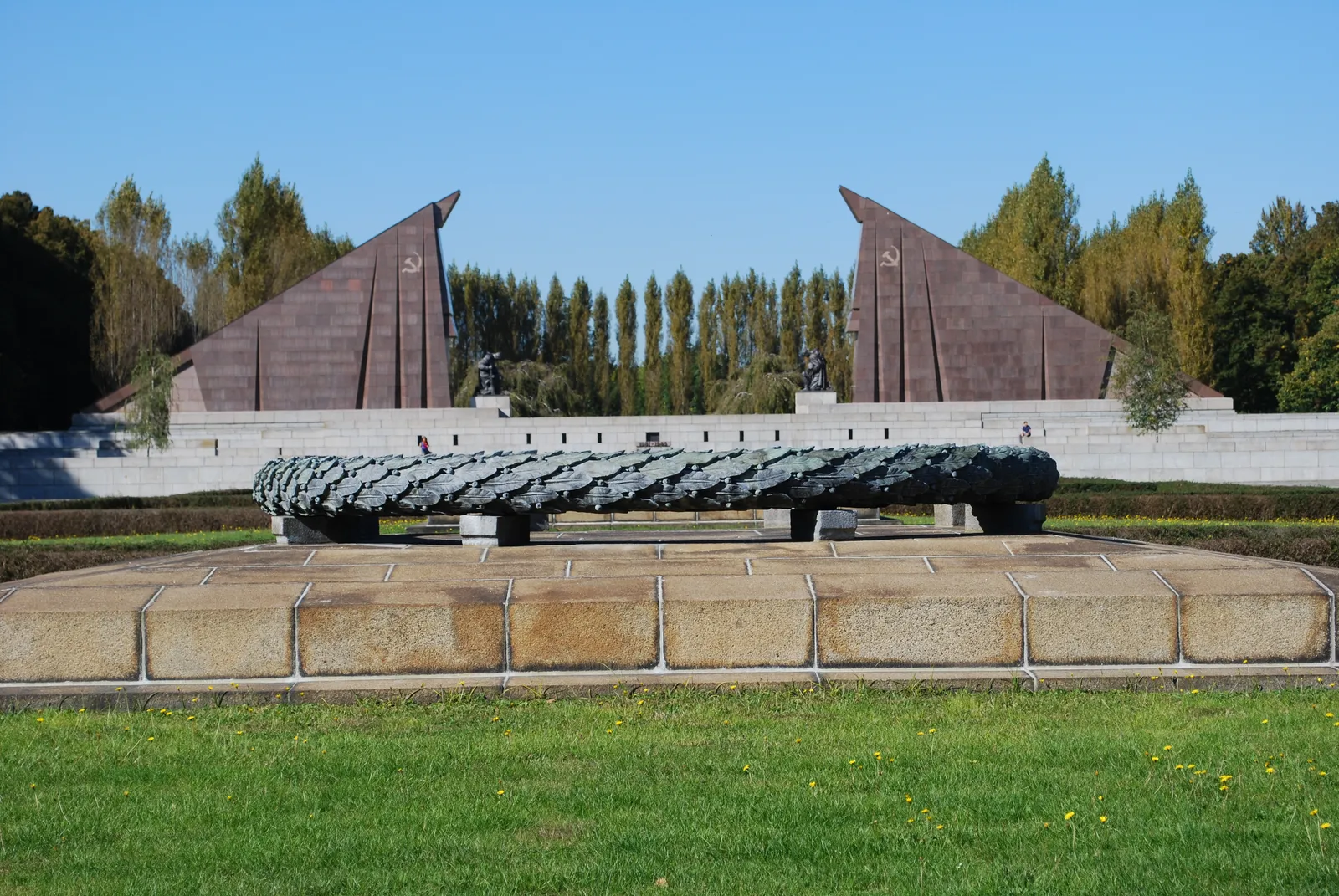 The Soviet War Memorial in Treptower Park, Berlin, with two enormous red granite flags angled toward each other over the central lawn and a row of clipped trees behind them under a blue sky