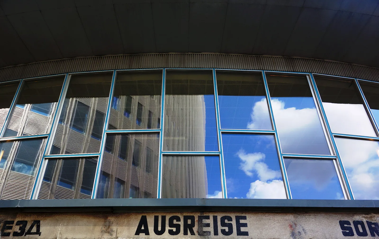 The curved glass front of the Tränenpalast in Berlin seen from below, reflecting sky and neighbouring buildings, with the original word AUSREISE lettered across the base