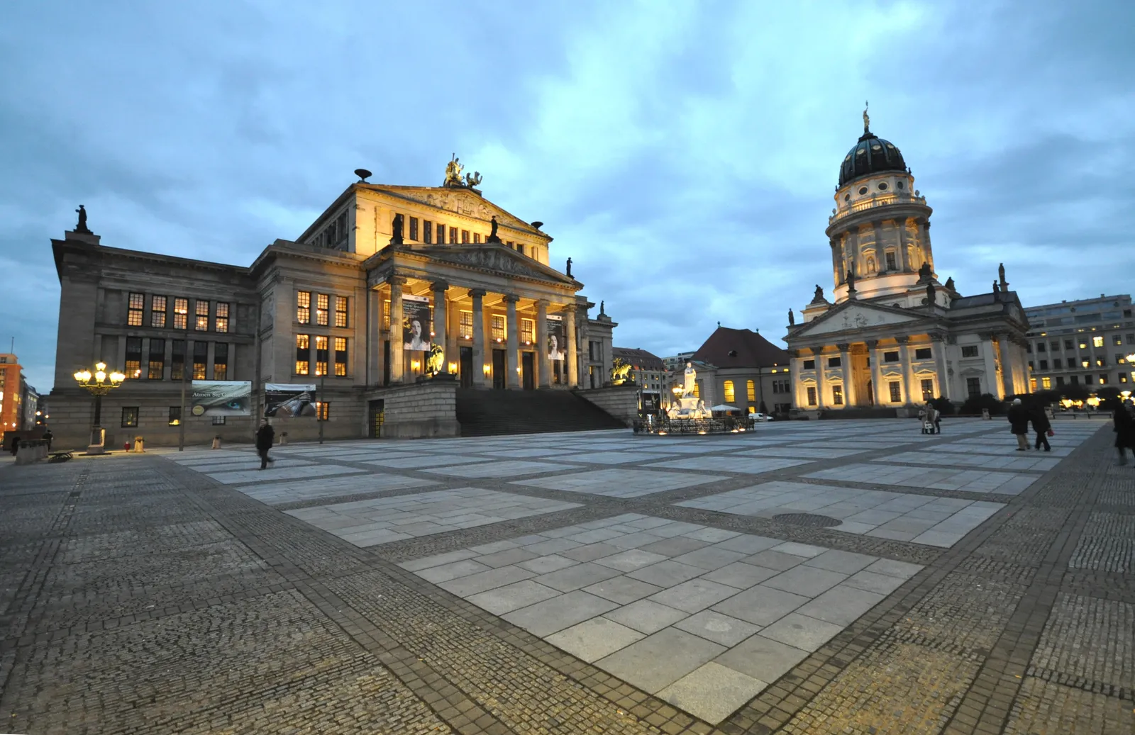 The Gendarmenmarkt in Berlin at dusk, with Schinkel's colonnaded Konzerthaus in the centre and the domed tower of the French Cathedral on the right, lit against a blue evening sky