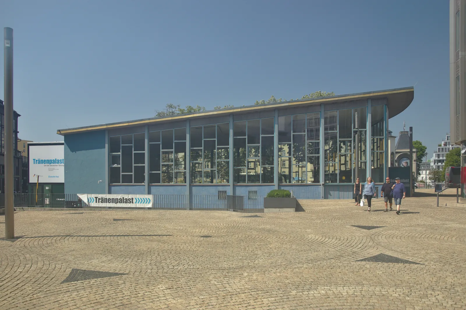 The Tränenpalast in Berlin, a low blue steel and glass pavilion beside Friedrichstraße station, with its name on a sign at the corner and people crossing the cobbled forecourt