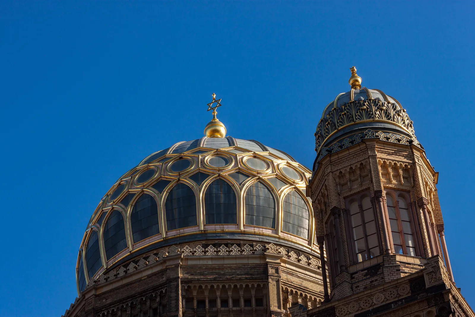 The gilded ribbed dome of the New Synagogue on Oranienburger Straße in Berlin against a clear blue sky, with its smaller flanking turret and ornate brickwork below