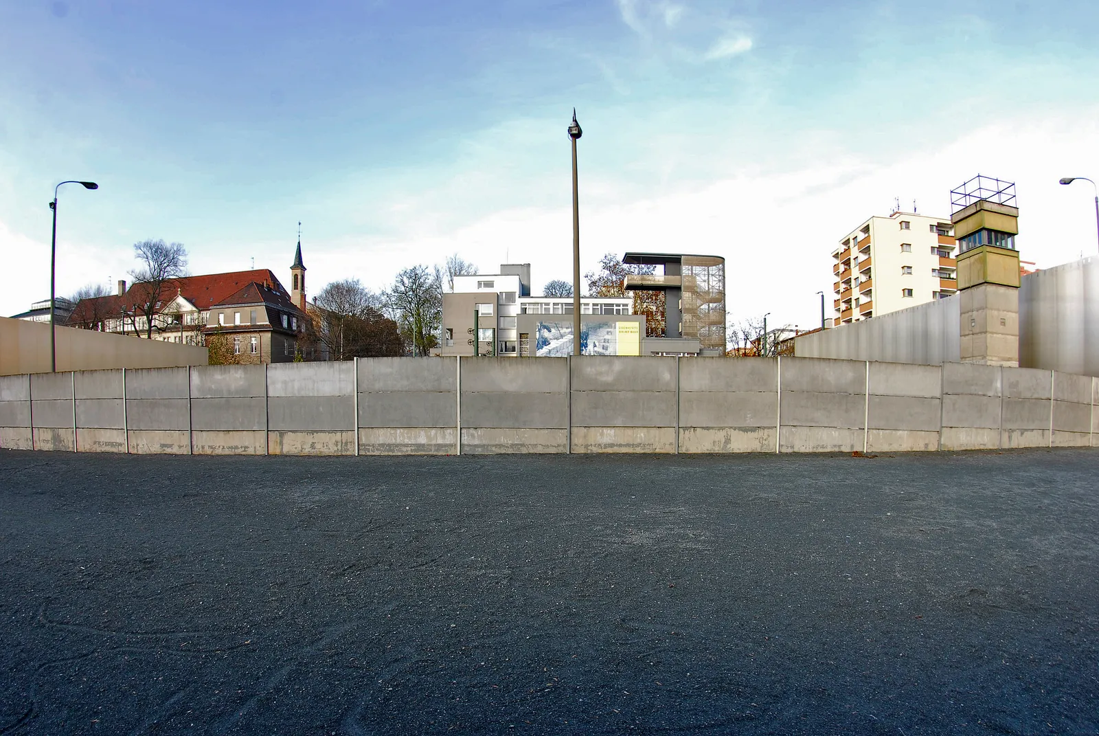 A preserved section of the Berlin Wall at the Bernauer Straße memorial, a run of grey concrete segments across the foreground with a surviving watchtower and apartment blocks behind it
