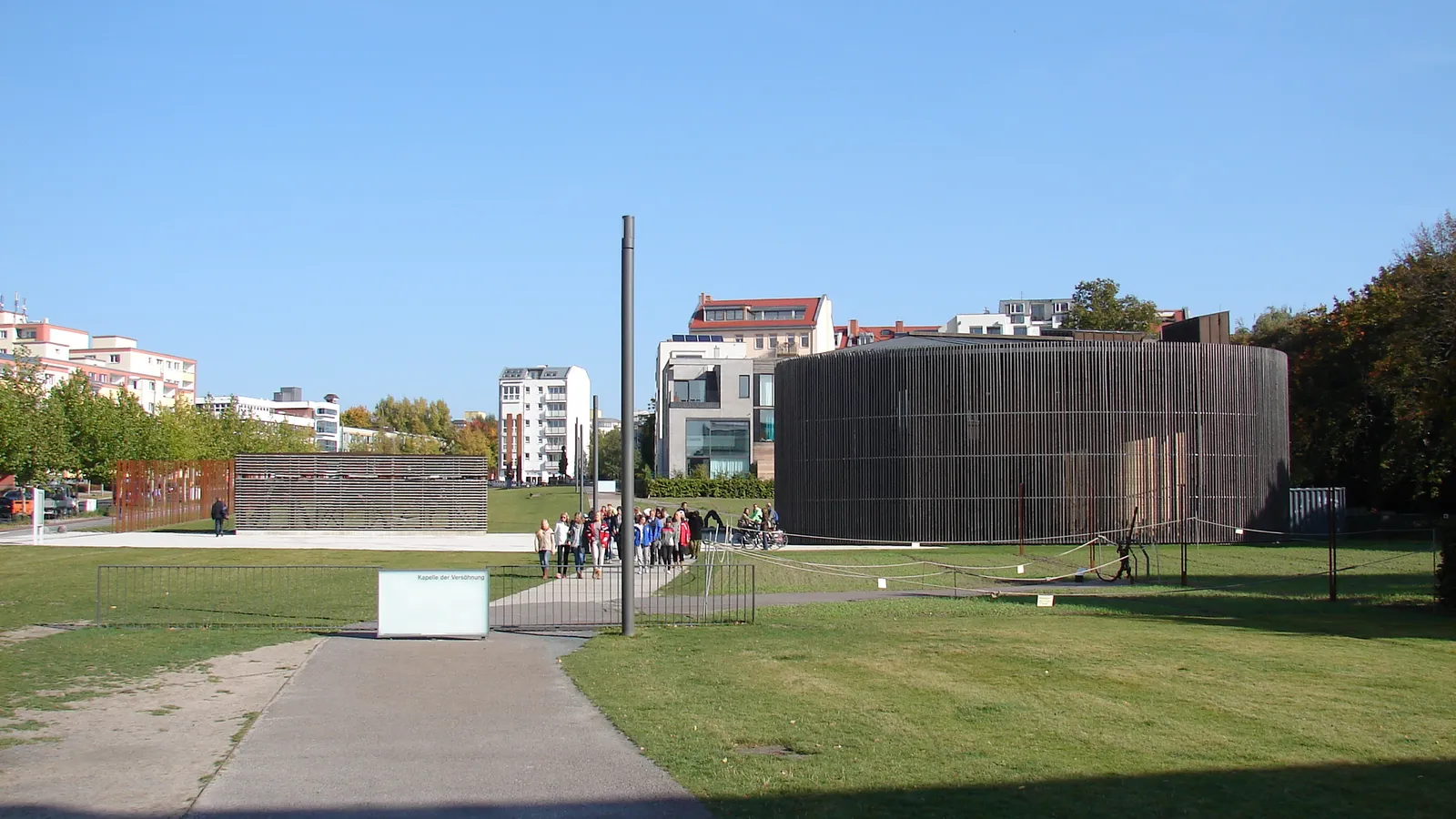 The oval Chapel of Reconciliation on the Berlin Wall memorial grounds, a low drum wrapped in vertical timber slats standing on open grass with wall segments and a group of visitors nearby