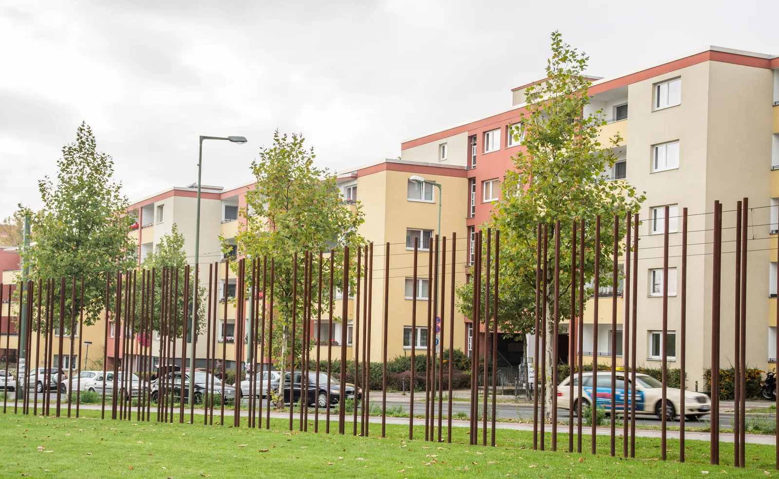 Rows of rusted steel posts marking the line of the vanished Berlin Wall on grass beside Bernauer Straße, with young trees and pastel post-war apartment blocks across the road