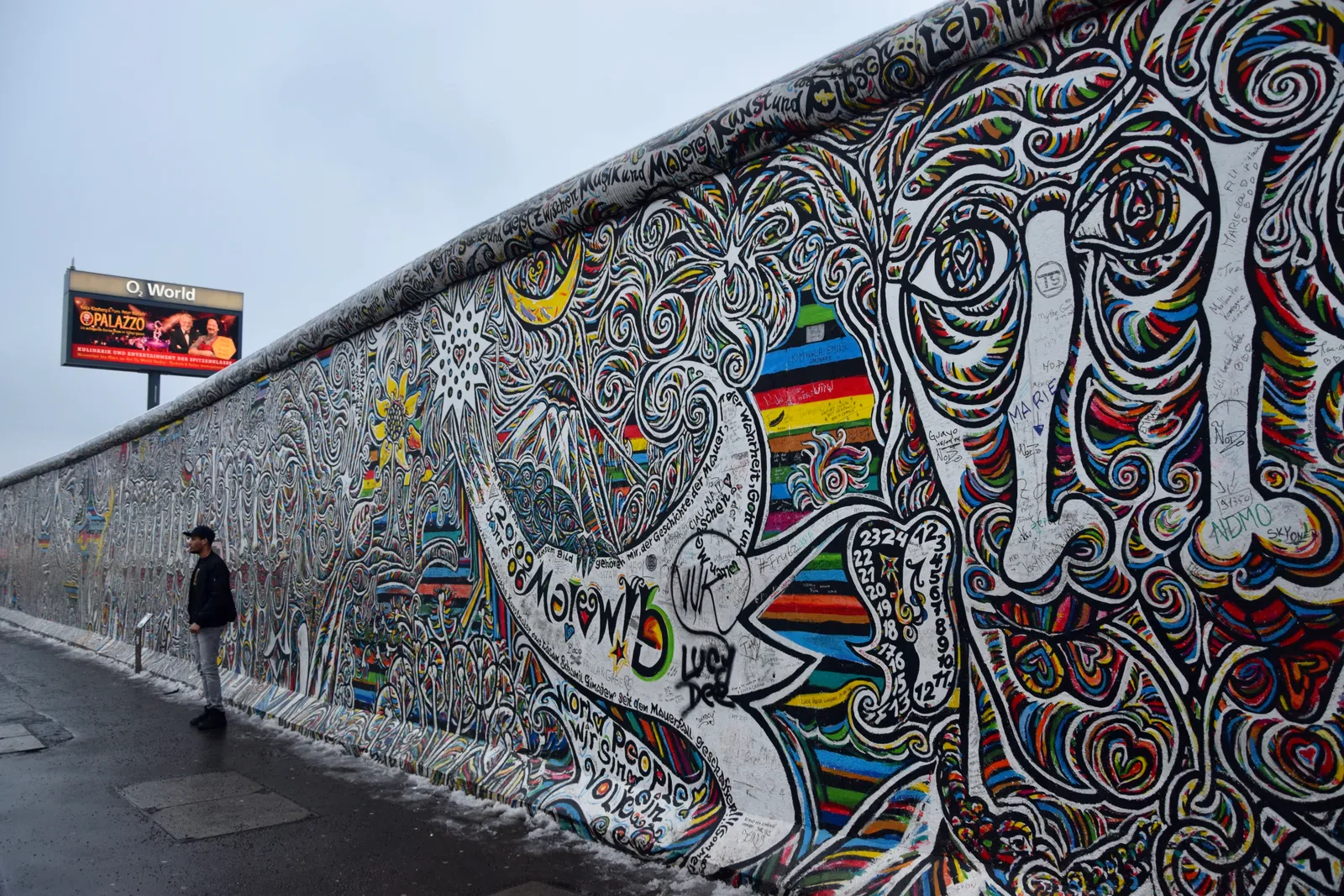 A long painted stretch of the East Side Gallery in Berlin running away down Mühlenstraße, covered end to end in bright murals, with a single person walking beside it