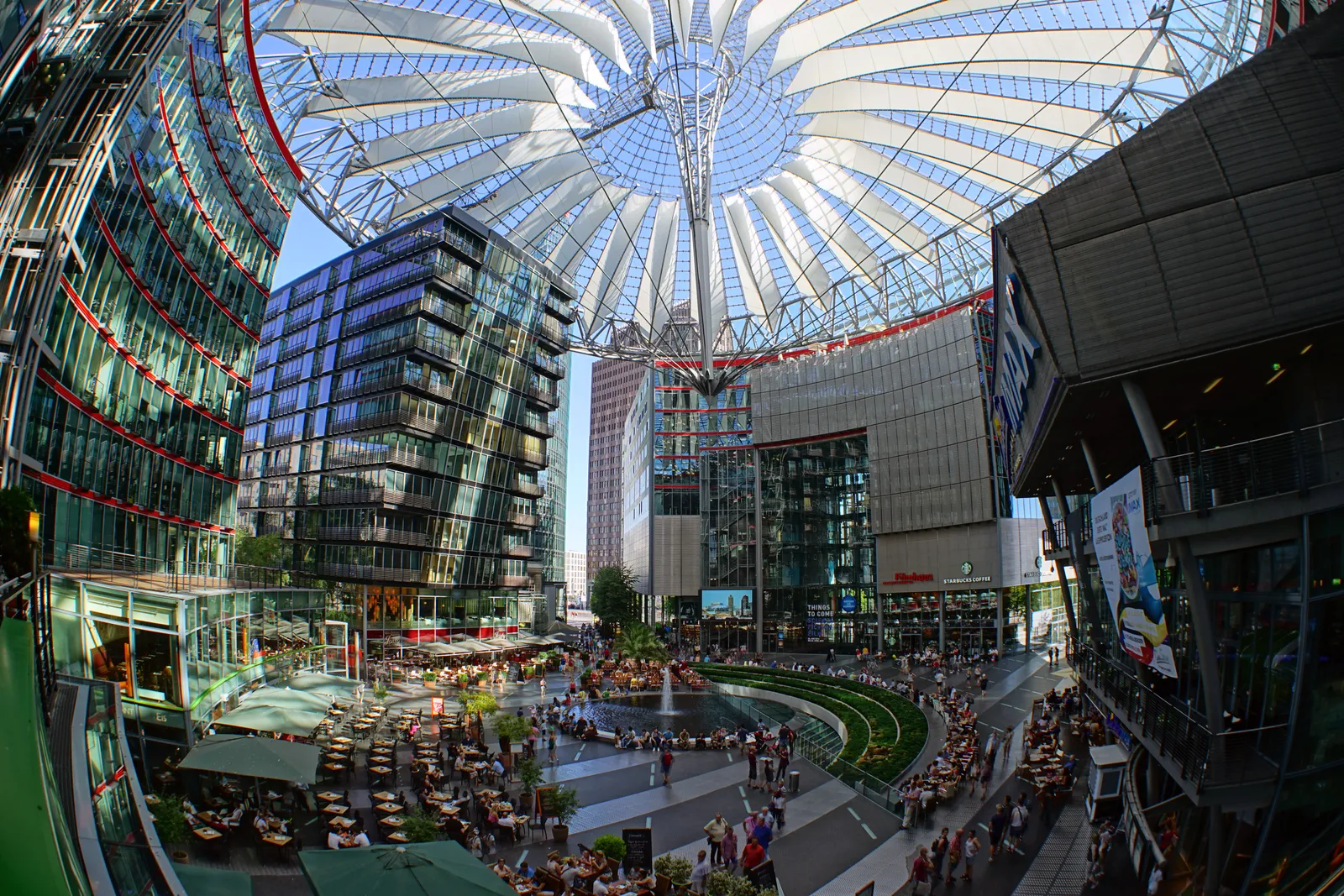 The interior court of the Sony Center at Potsdamer Platz in Berlin, ringed by glass office fronts under a tent-like fabric and steel roof, with café tables and people crossing the paving below