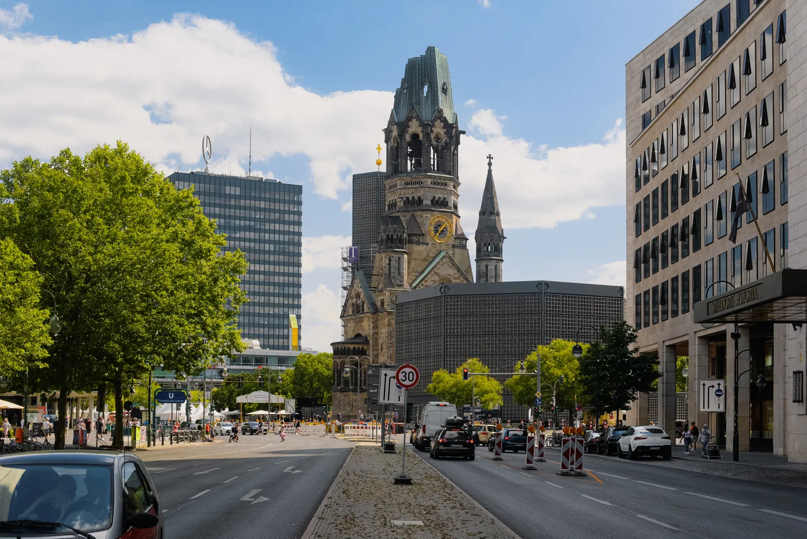 The Kaiser Wilhelm Memorial Church in Berlin seen down a tree-lined street, its jagged bomb-damaged tower rising above the traffic with the low modern church beside it