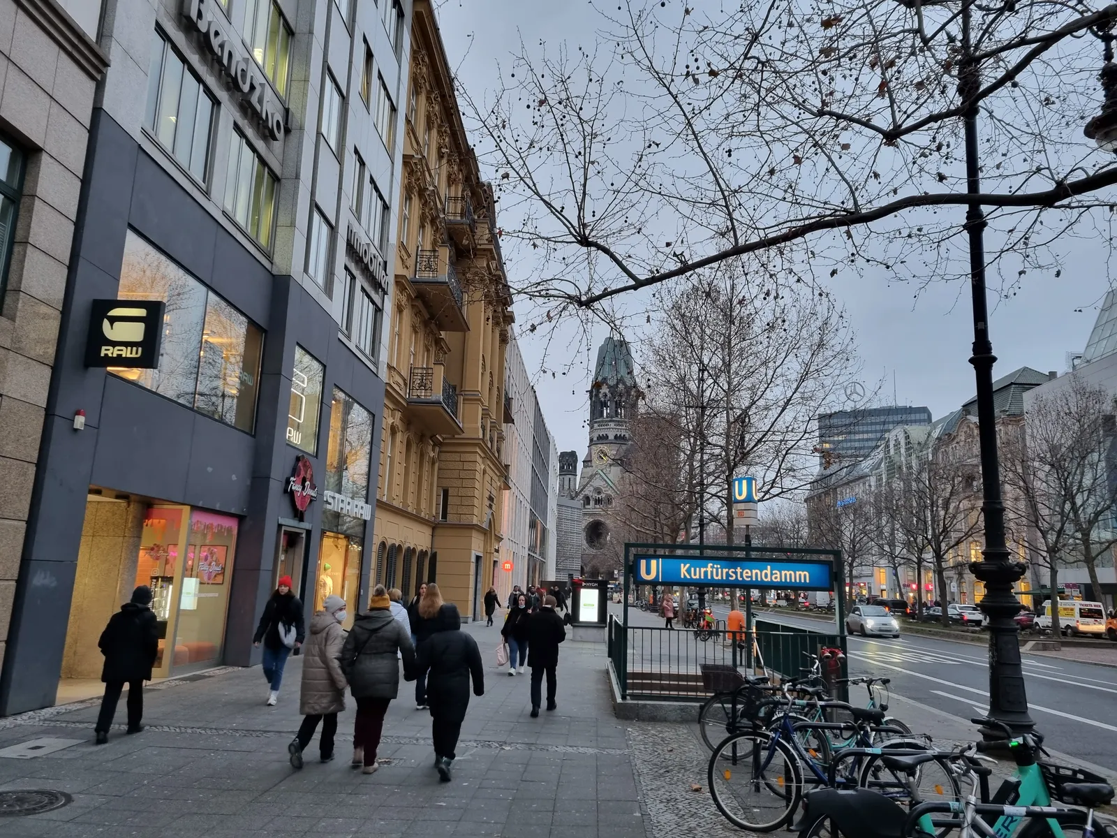 A pavement on the Kurfürstendamm in Berlin on a winter afternoon, with shopfronts on the left, a green U-Bahn entrance sign reading Kurfürstendamm, and the Memorial Church tower closing the view