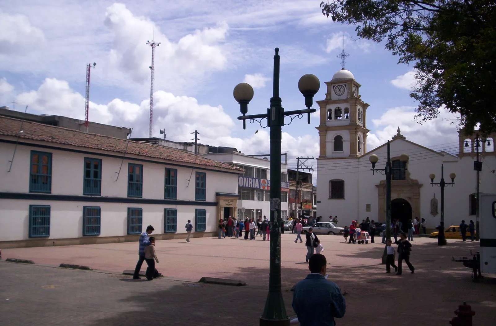 The founding plaza of Fontibón in Bogotá, an open paved square with people crossing it, low colonial buildings along one side and the white church with its clock tower on the far corner