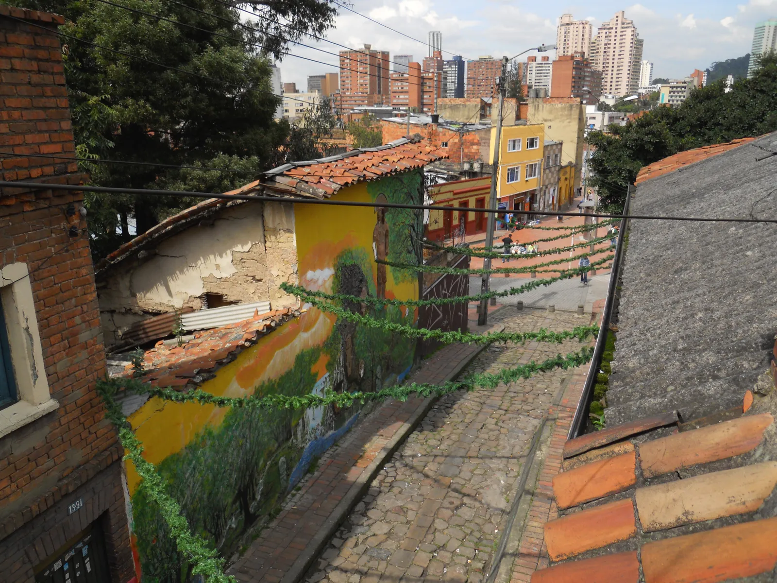 The cobbled Calle del Embudo in Bogotá seen from above, a narrow lane dropping between painted low houses with tiled roofs and the city's towers on the skyline beyond