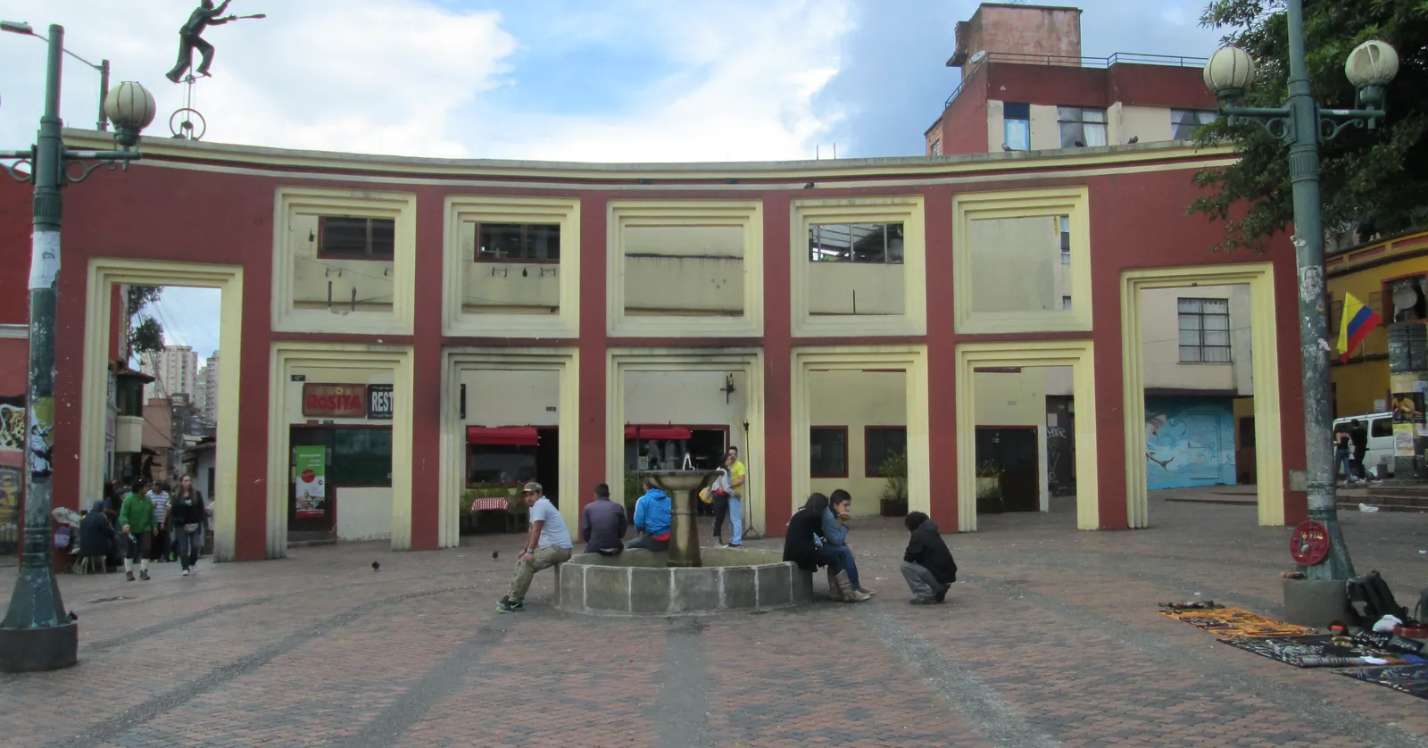 The Chorro de Quevedo square in Bogotá, a small paved plaza with a stone fountain in the middle and a curved red and cream building of shopfronts closing off one side