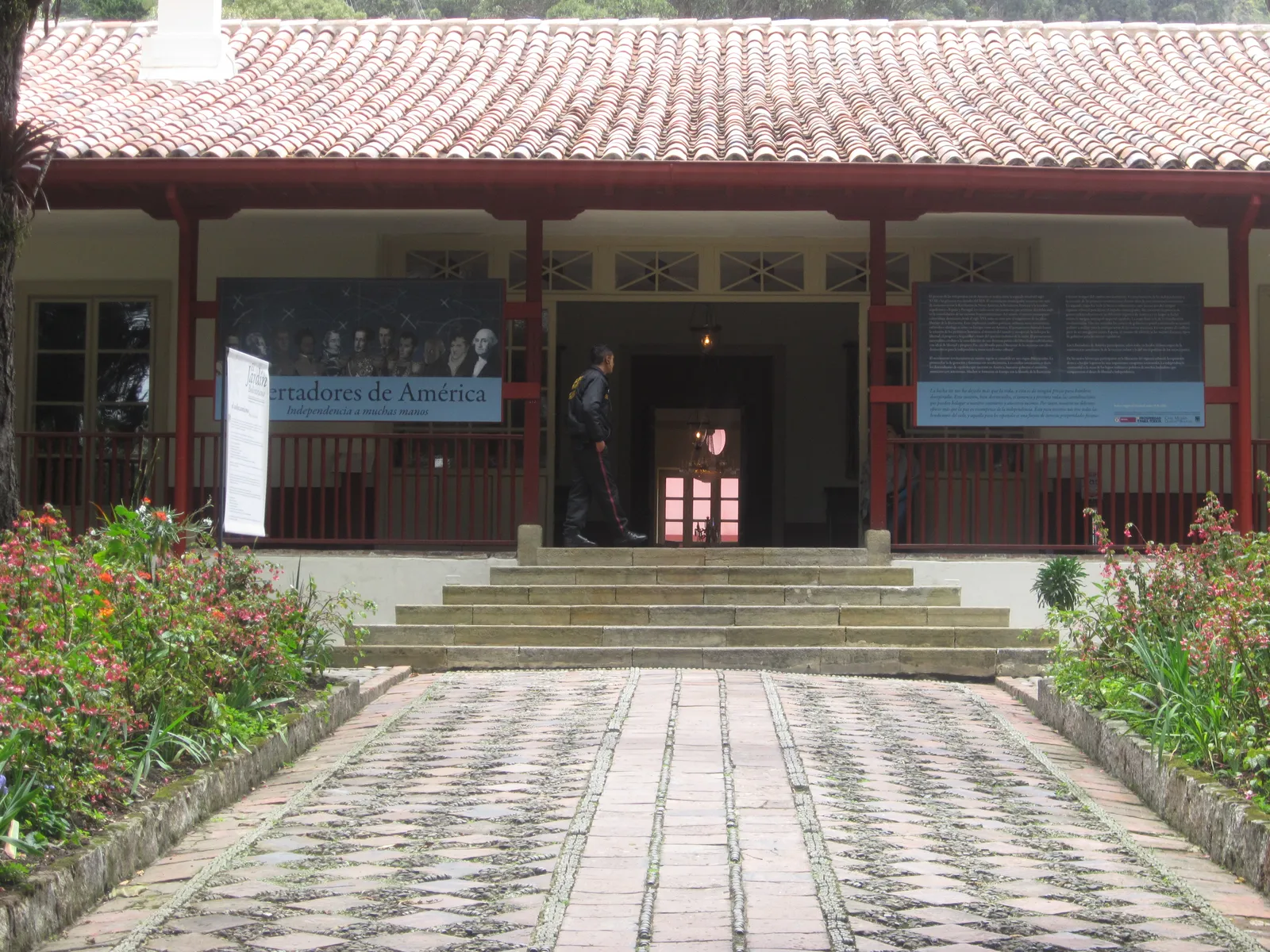 The front of the Quinta de Bolívar house museum in Bogotá, a single-storey white house with red posts holding up a tiled veranda roof, reached by a brick path through a flower garden