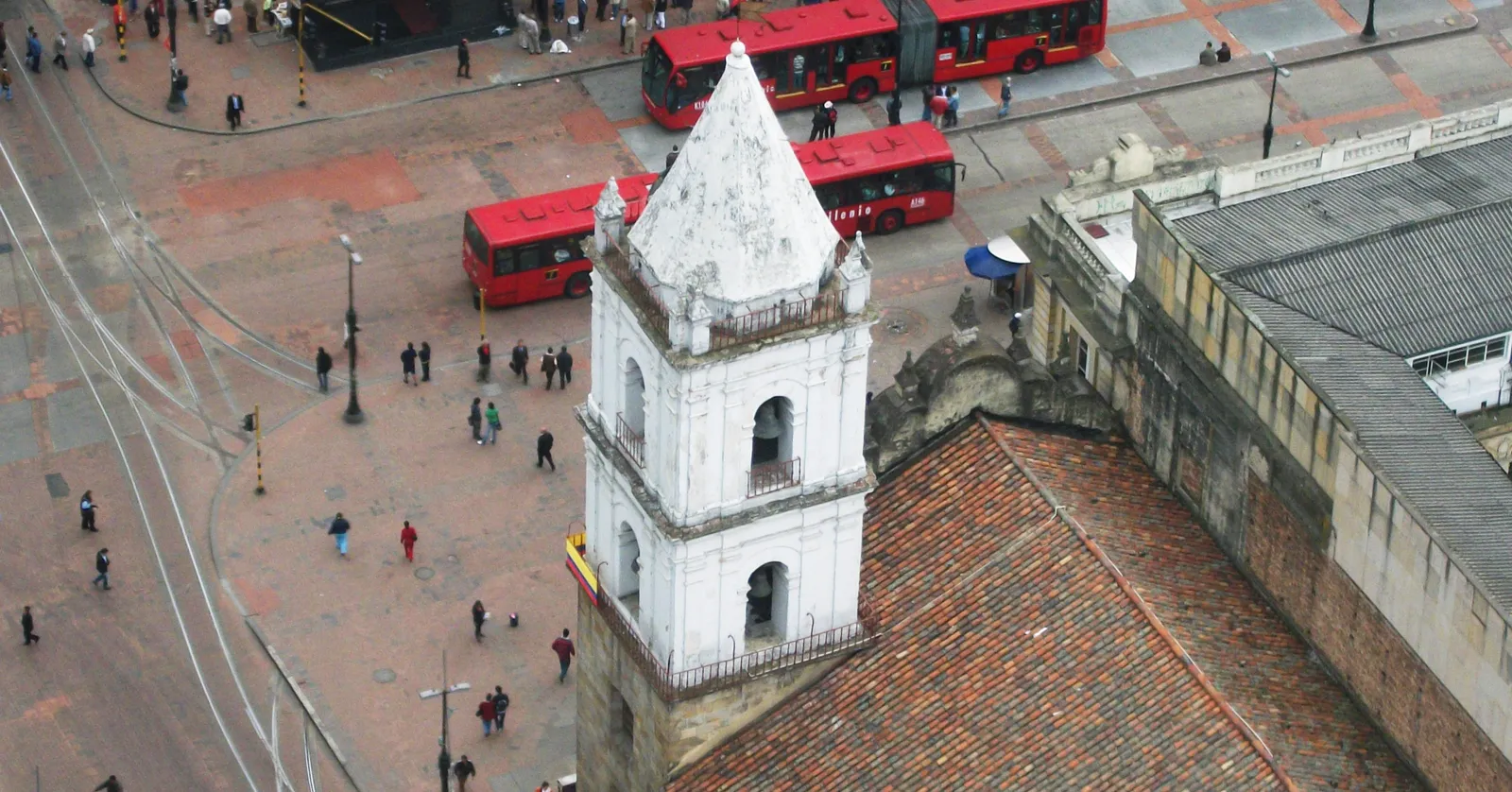 Looking down on the white bell tower and tiled roof of the San Francisco church in Bogotá, with Avenida Jiménez below it carrying red articulated buses and crowds of pedestrians