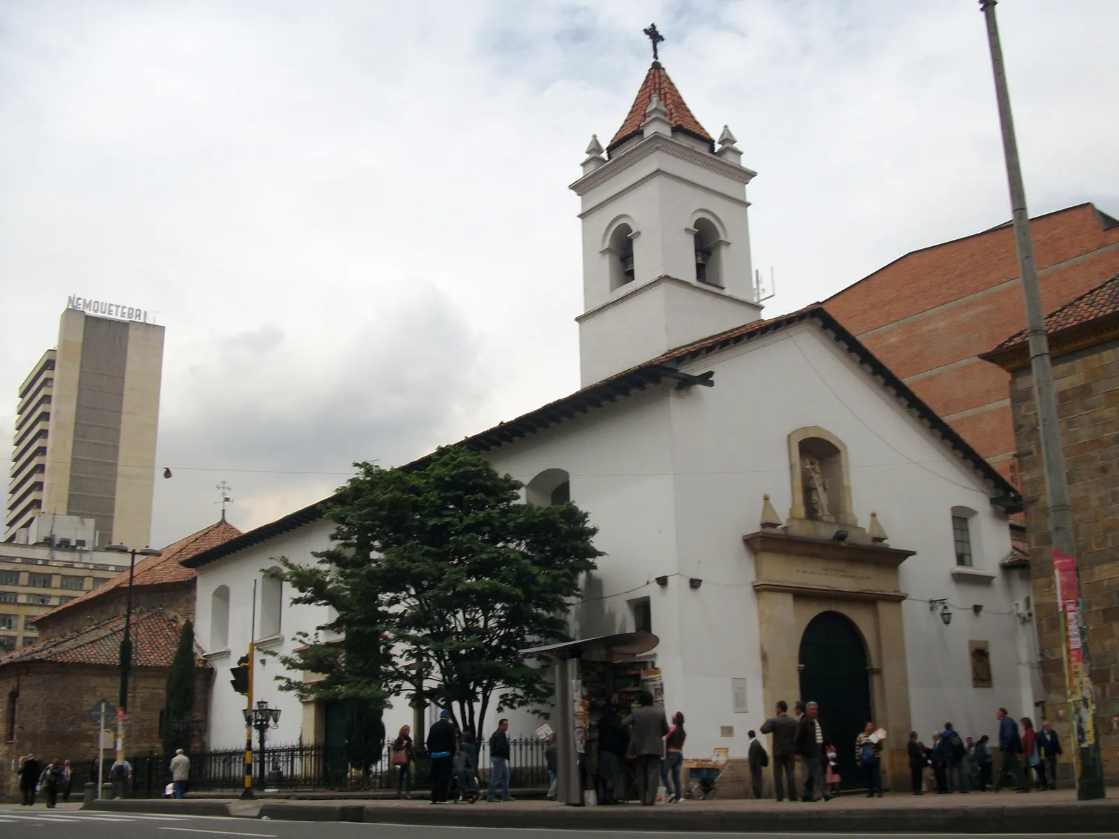 The whitewashed Iglesia de la Veracruz in Bogotá seen from the street corner, with a square bell tower under a tiled roof and people waiting outside its arched doorway