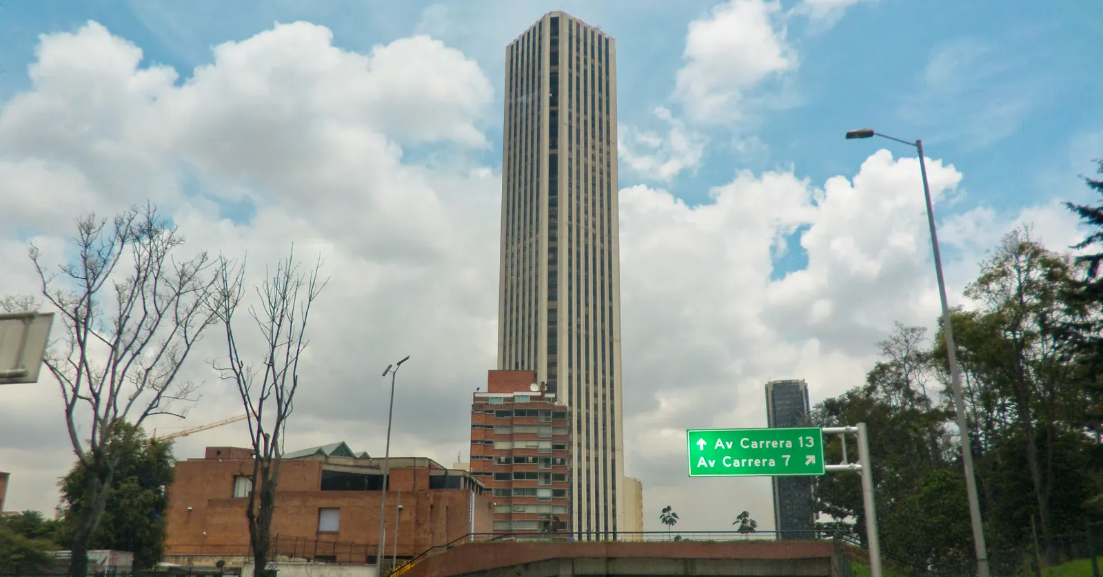 The Torre Colpatria in Bogotá, a plain slab-sided skyscraper of more than fifty floors rising above low brick buildings and road signs under a cloudy blue sky