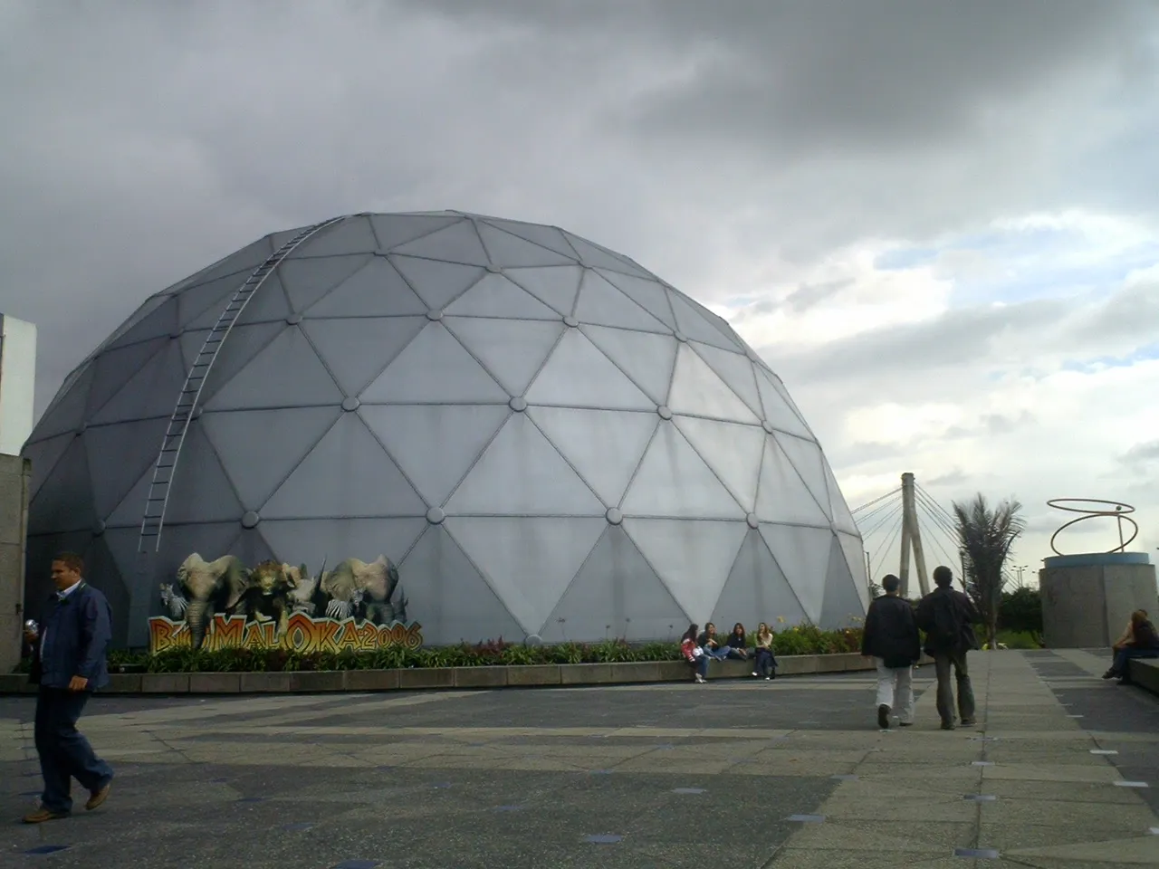 The grey geodesic dome housing Maloka's cinema in Bogotá, rising from a paved forecourt with people crossing in front of it under a heavy sky