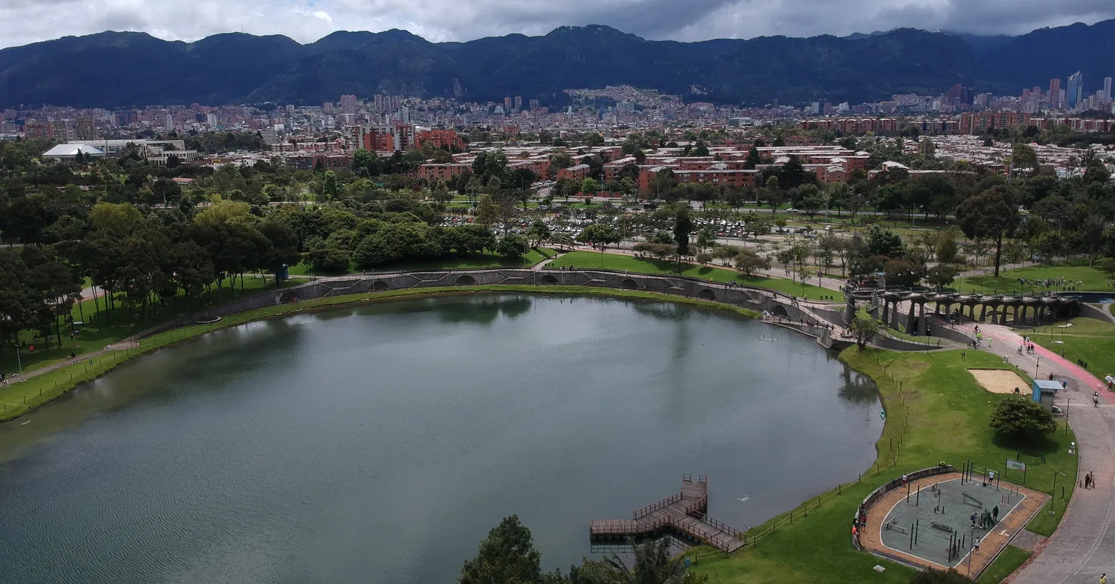 Aerial view of the boating lake in Simón Bolívar park in Bogotá, ringed by trees and paths, with apartment blocks and the eastern mountains behind it